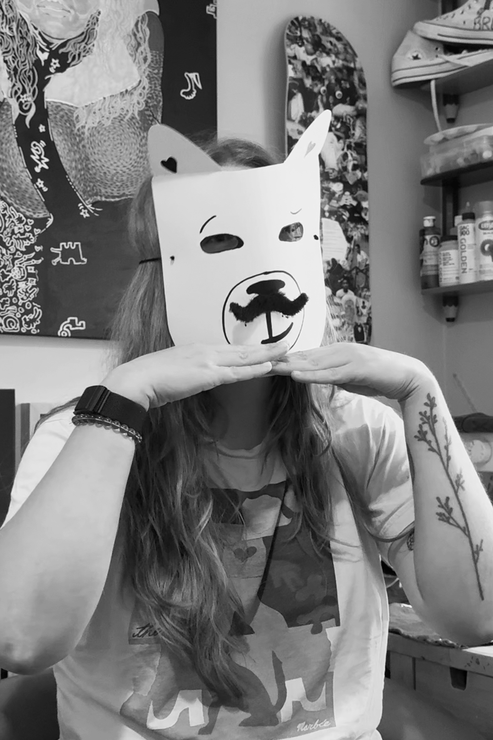 Person with long hair wearing a T-shirt, holding a mask with a dog face over their face, with their hands under their chin. The background shows artwork and shelves with bottles.