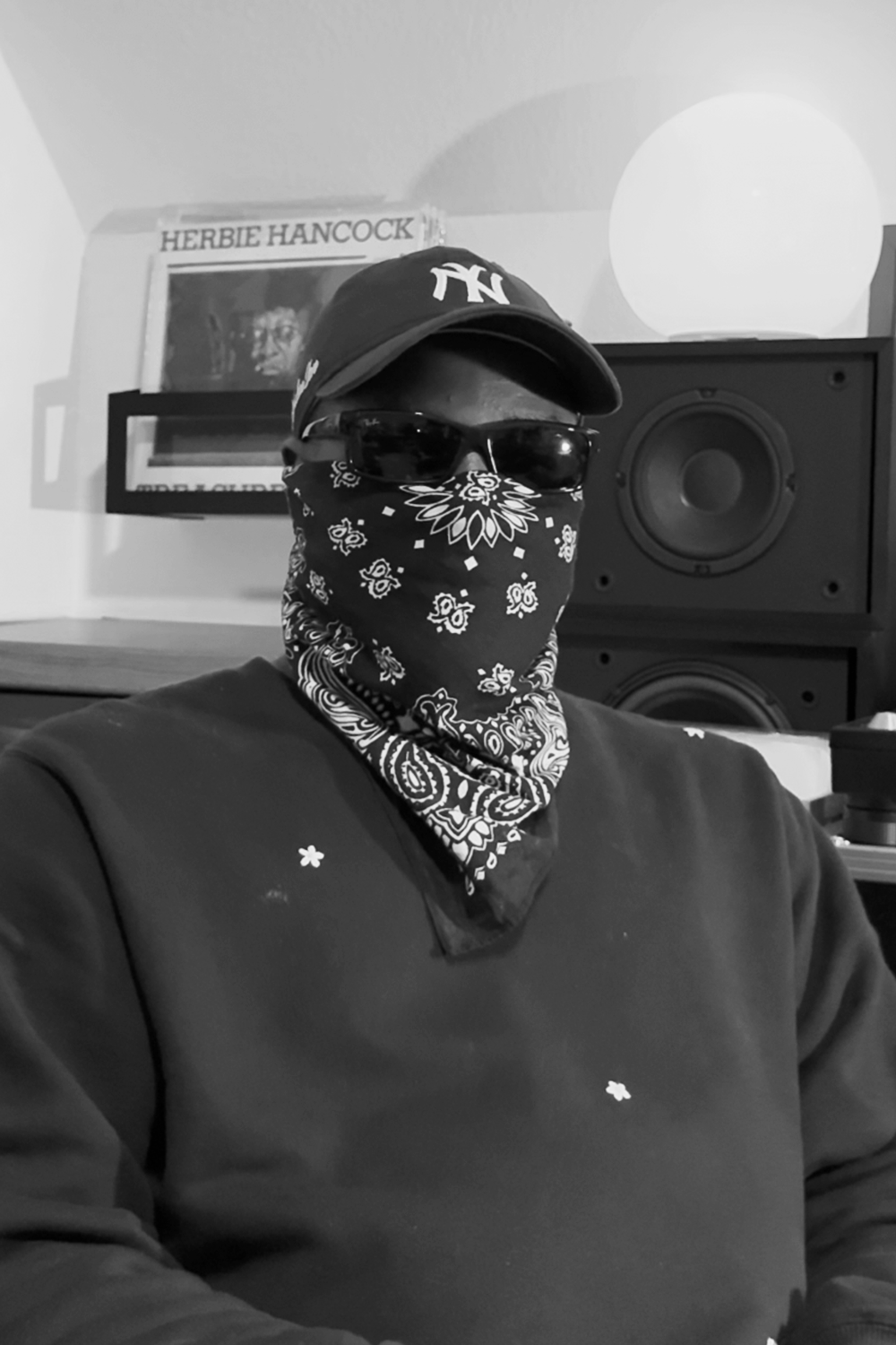 Person wearing sunglasses, a cap, and a bandana covering the face, sitting in a room with a bookshelf, record, and speaker visible in the background.