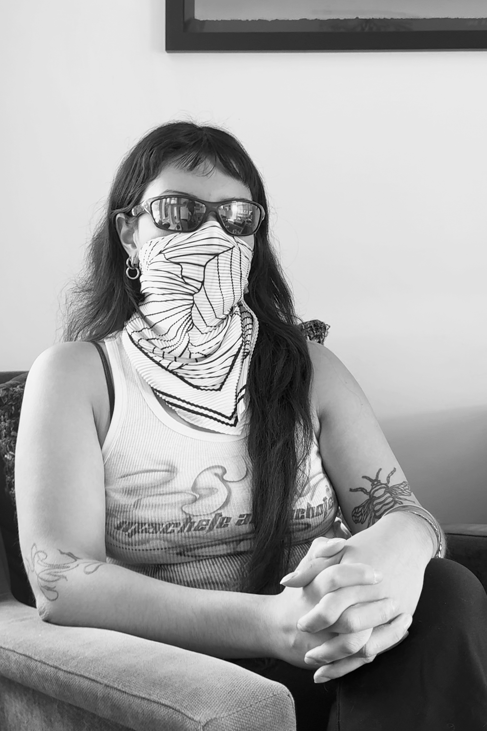 Woman wearing sunglasses and a bandana covering her face, sitting with her hands clasped on her lap in a casual setting.