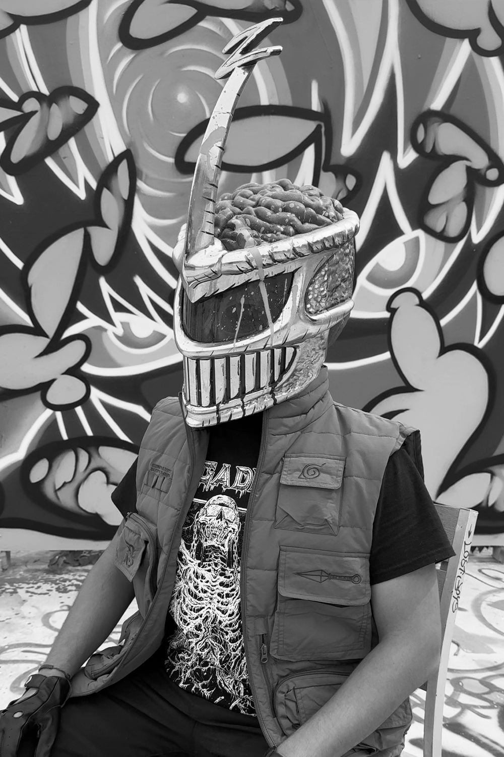 Person wearing a skull-shaped metal sculpture mask with a brain-like top, sitting in front of a colorful graffiti background.