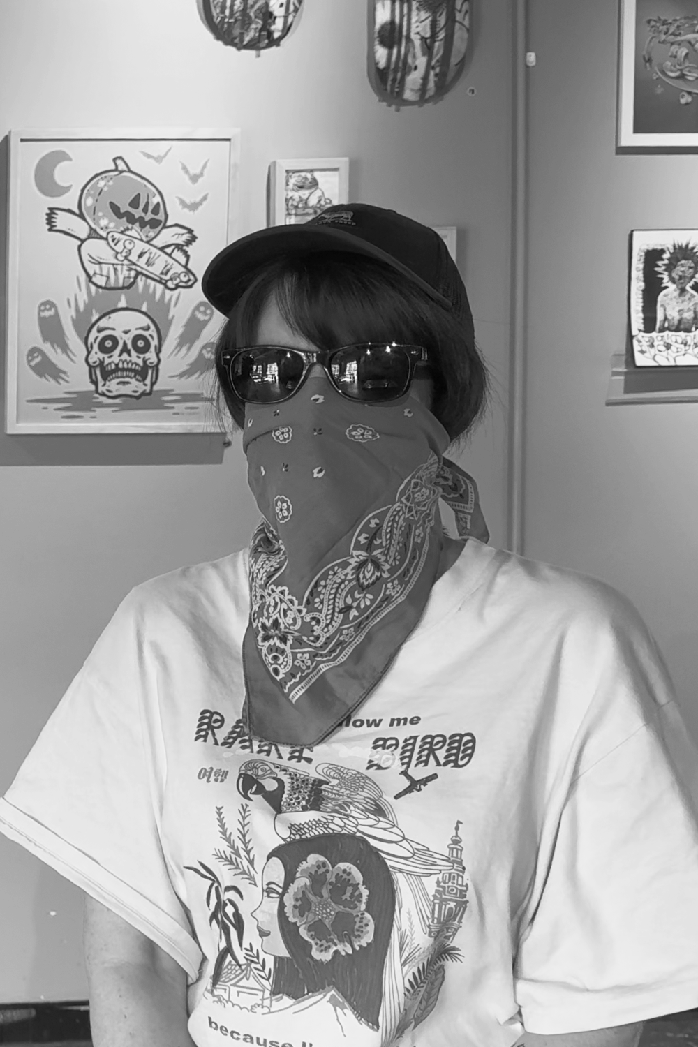 Person wearing glasses, a bandana covering face, a cap, and a graphic T-shirt, standing in front of Halloween-themed artwork.