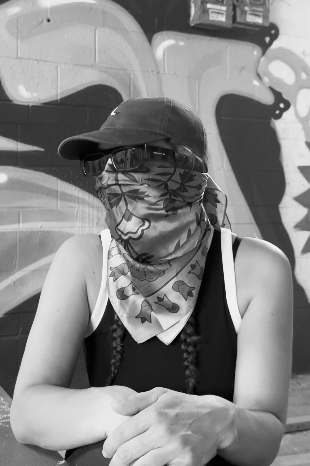 Person wearing a baseball cap, sunglasses, and a bandana covering their lower face, sitting in front of a mural on a brick wall.