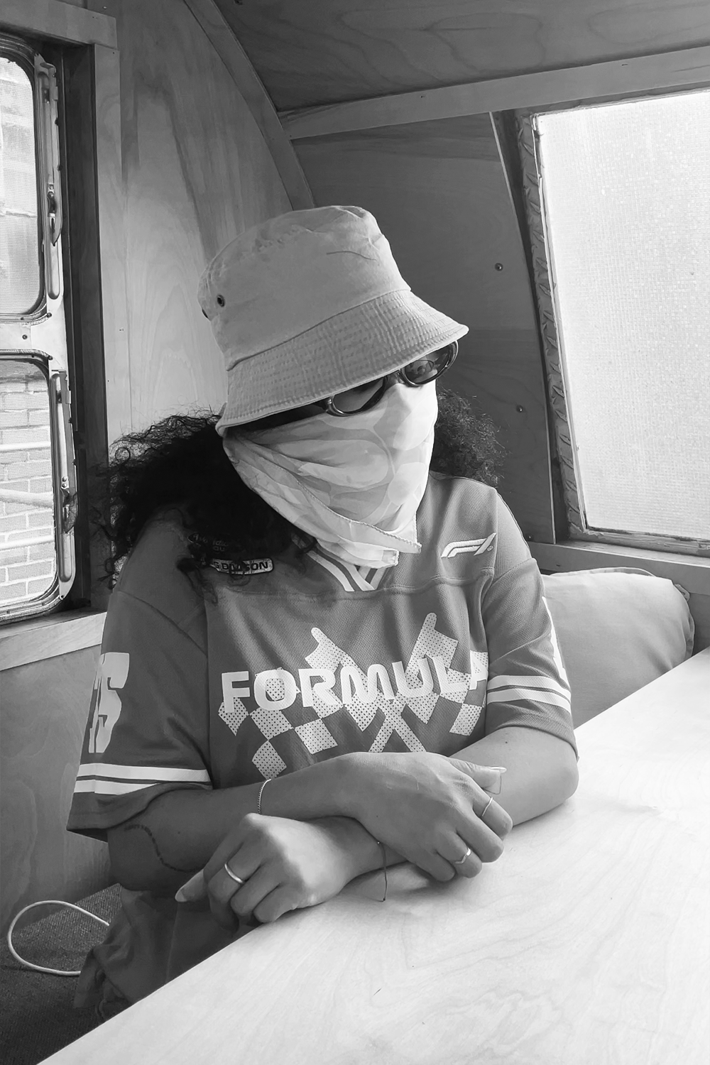 A woman wearing a bucket hat, sunglasses, a face mask, and a Formula 1 shirt sitting at a table inside a vehicle or wooden structure with two small windows.