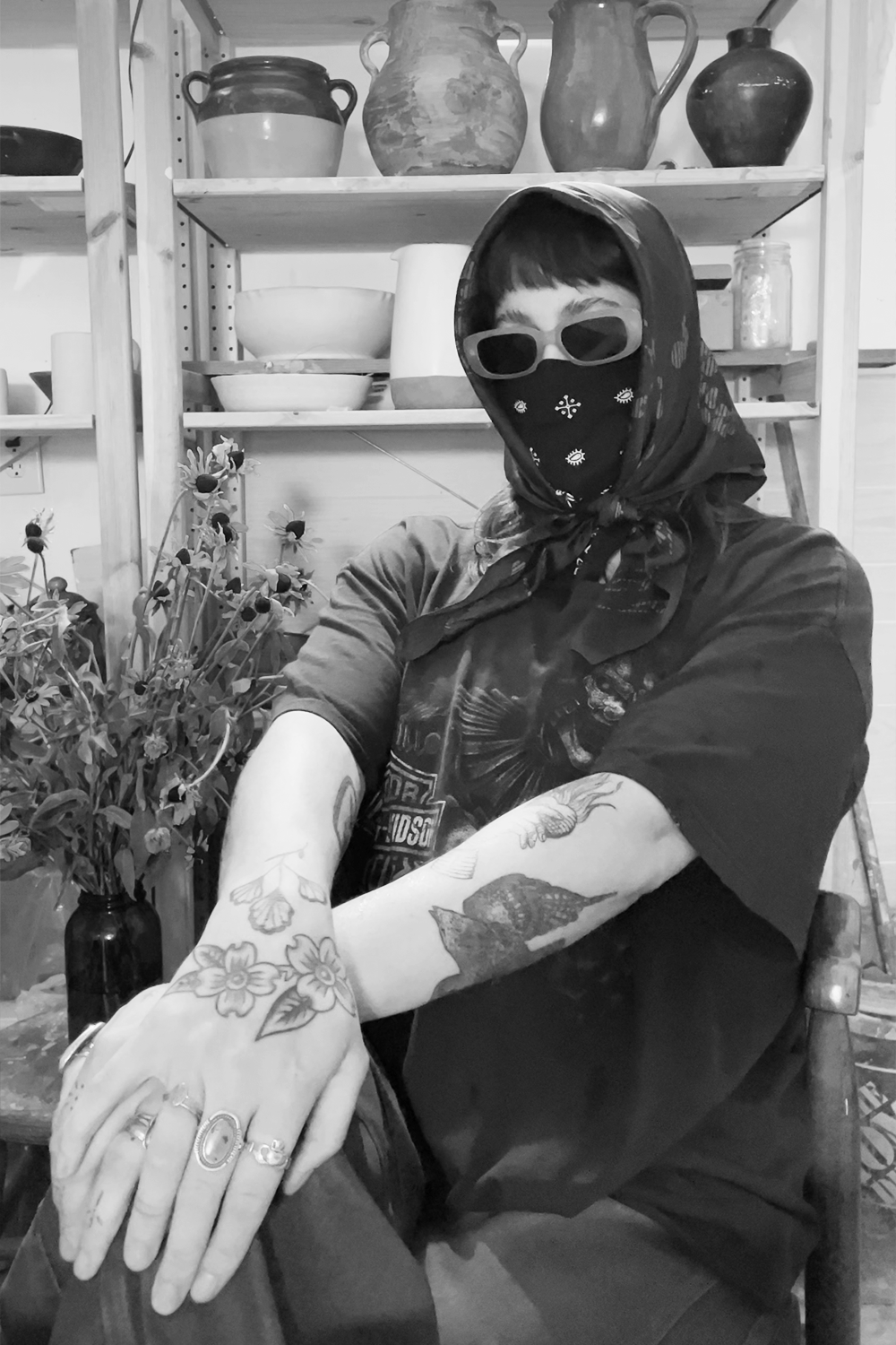 A person with tattoos on their arms, wearing sunglasses, a bandana covering their face, and a hooded shirt, sitting beside a vase of flowers in a room with shelves of pottery and jars in the background.