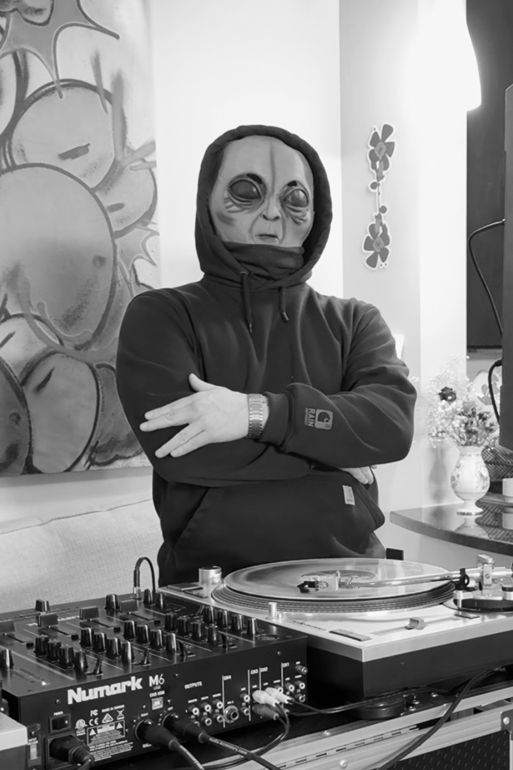 Person wearing a mask with an alien face design, dressed in a hoodie, standing behind DJ equipment in a room with wall art and a vase with flowers.