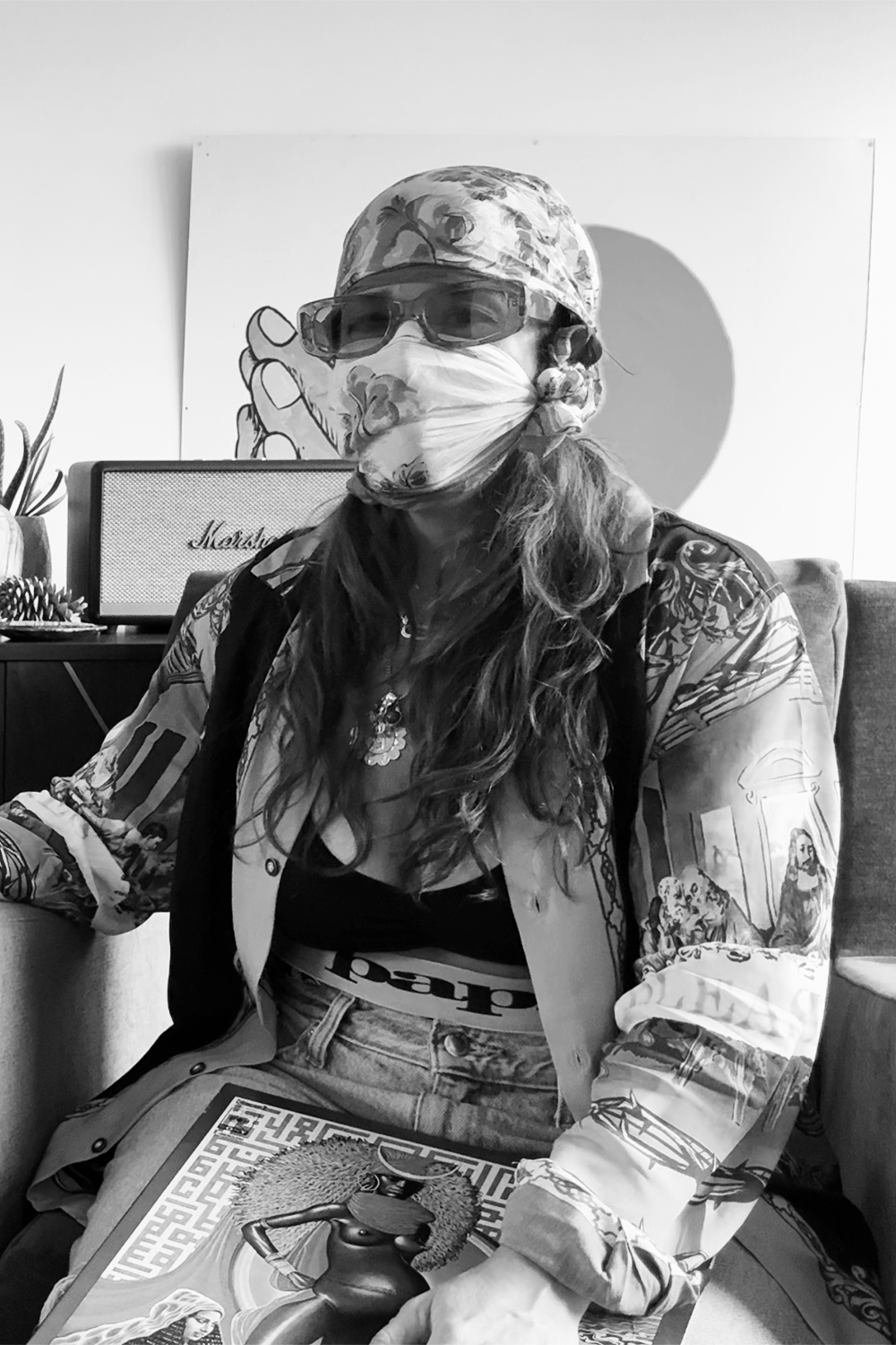 A person with long wavy hair wearing a patterned bucket hat, sunglasses, a face mask, a floral jacket, and a necklace, seated indoors with a magazine on their lap, and a painting of a person with hands clasped in the background.