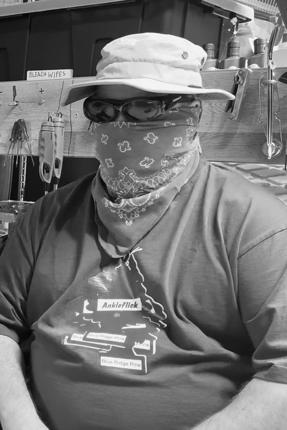 A person wearing a hat, sunglasses, bandana covering their face, and a t-shirt with a tree diagram, sitting in a workshop with tools and supplies in the background.