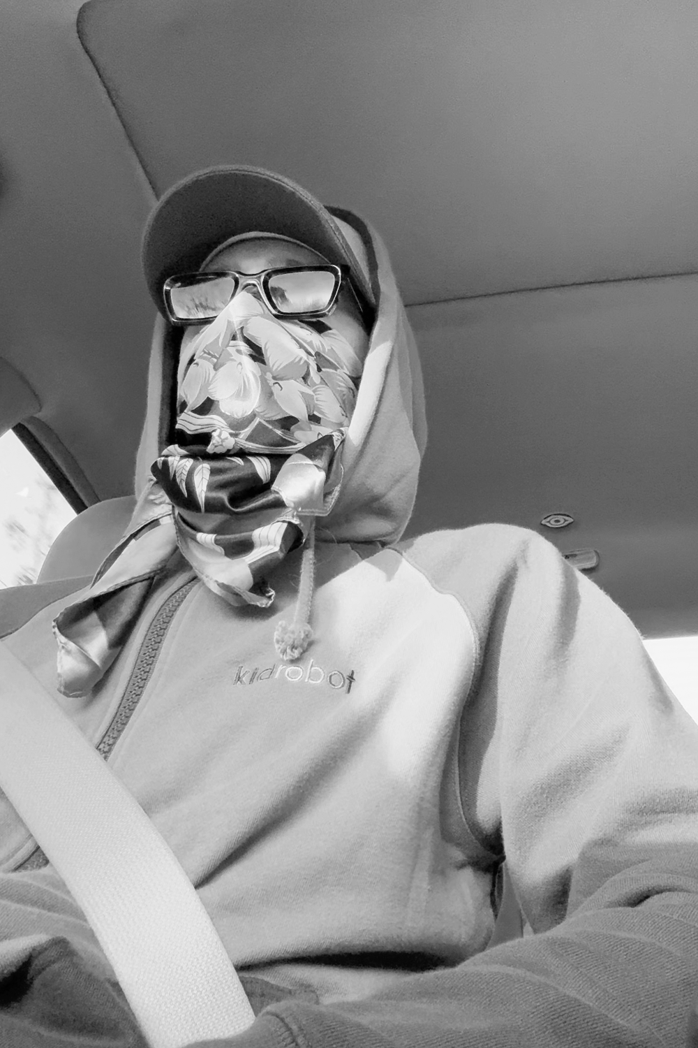 Person wearing a hoodie, scarf, glasses, and cap, sitting in a car.