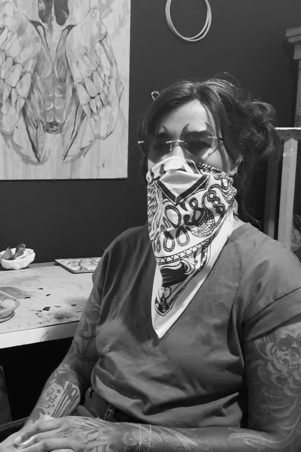 Person with tattoos, wearing sunglasses and a bandana covering face, sitting at a table in a tattoo studio.