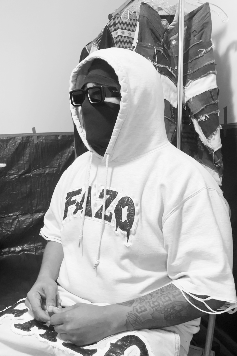 Man dressed in a white hoodie with the word 'FALLO' on it, wearing dark sunglasses and a face covering, seated in a room with hanging clothing and plastic-covered objects in the background.