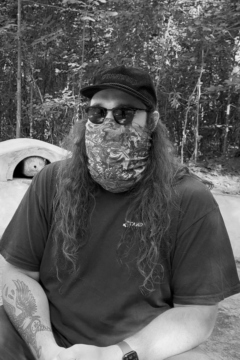 A person with long hair wearing sunglasses, a patterned face covering, a black cap, and a T-shirt sits outdoors in a wooded area.