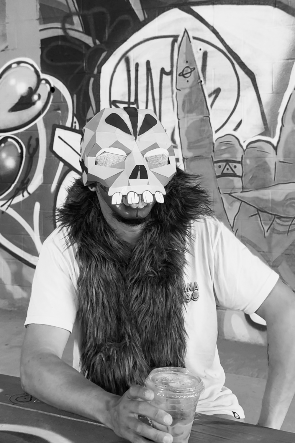 Person wearing a cartoonish skull mask with exaggerated features, a faux fur scarf, and holding a cup, sitting in front of a graffiti mural with drawings of planets and a large hand making a peace sign.