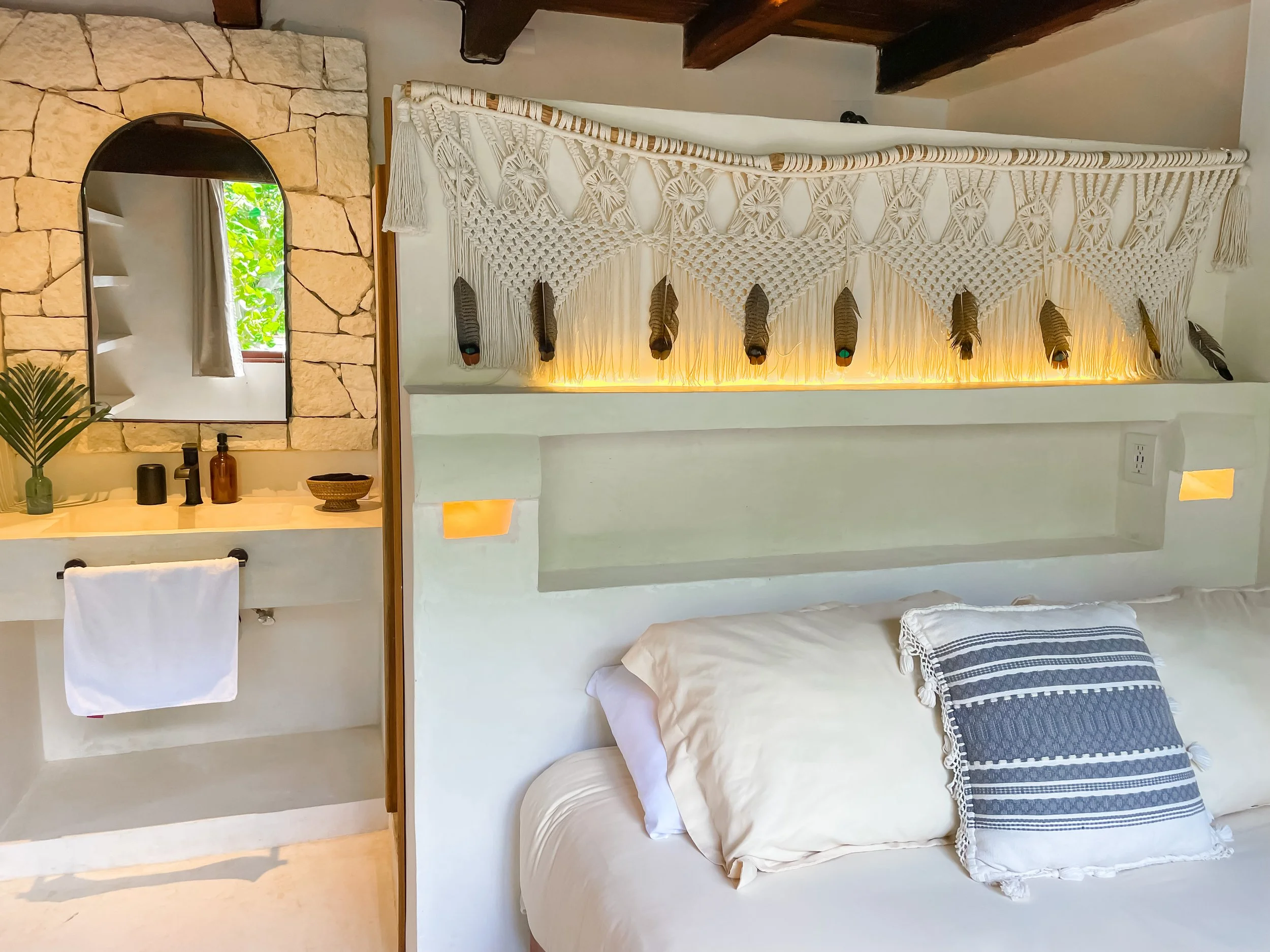 A cozy bedroom with a bed featuring white pillows and a decorative blue and white pillow, a built-in shelf with warm lighting, a hammock with feathers hanging above the bed, and a bathroom vanity area with a mirror, vases, soap dispensers, and a towel.