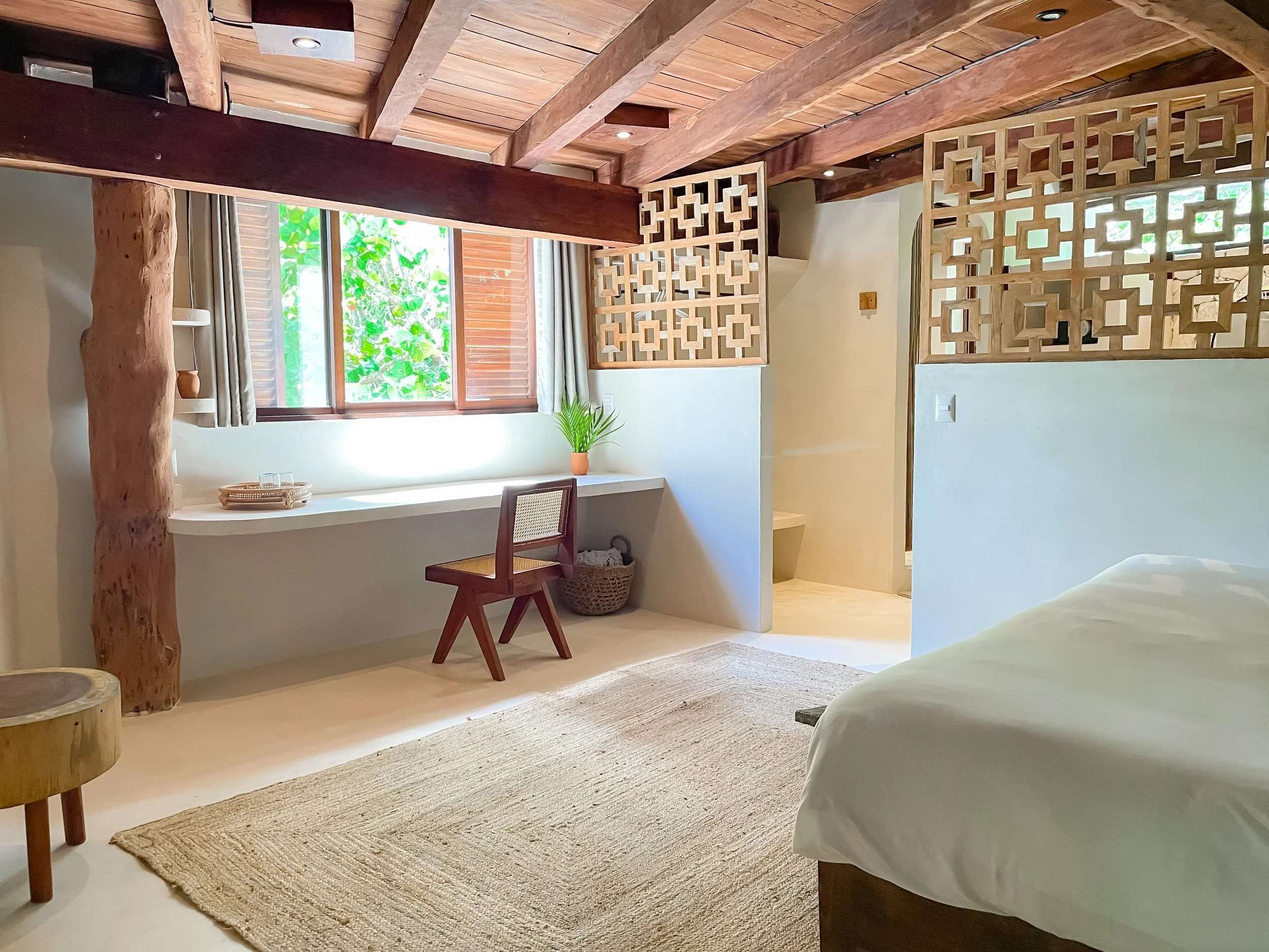 A cozy bedroom with a wooden ceiling, large window with curtains, and a bed with white linens. There's a small desk with a chair near the window, a potted plant on the windowsill, and intricate wooden room dividers. The room has a neutral color palette with natural wood accents and a beige rug on the floor.