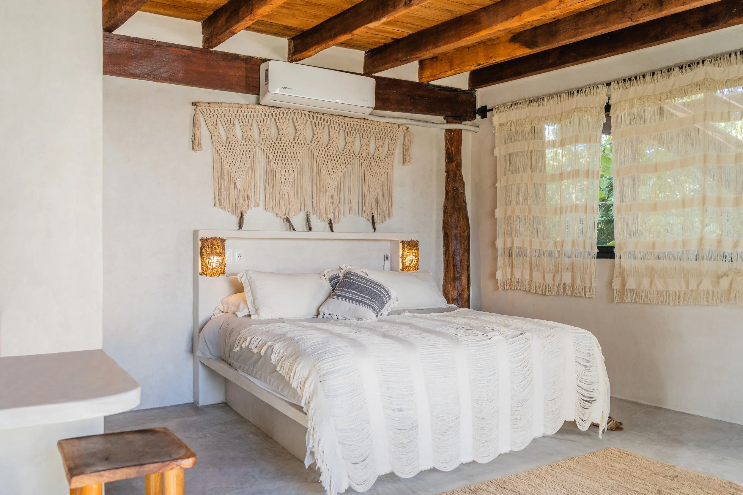 A cozy bedroom with a white bed, decorative pillows, and wood accents. The room features a macrame wall hanging above the bed, woven curtains on the windows, and exposed wooden ceiling beams.