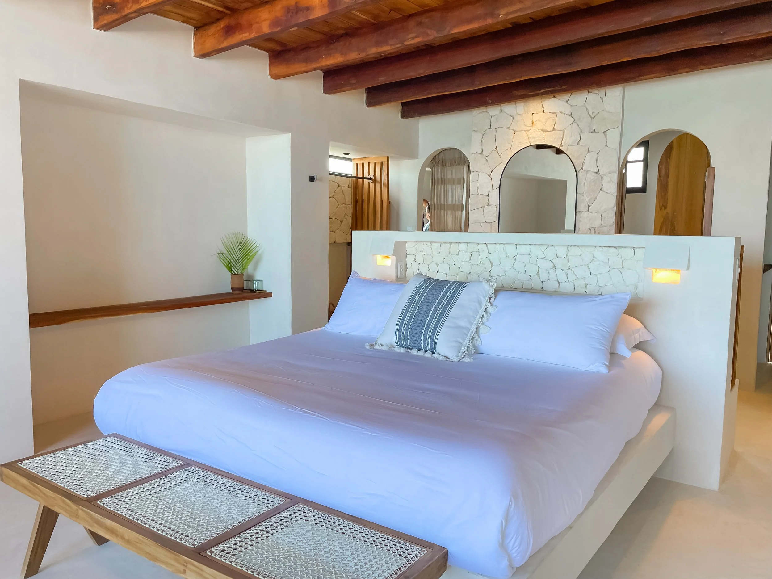 A bedroom with a white modern bed, a decorative pillow, and a wooden bench at the foot of the bed. The headboard doubles as a partial divider wall. The room has exposed wooden beams on the ceiling, a built-in shelf with a small plant, and arched doorways with stone accents.