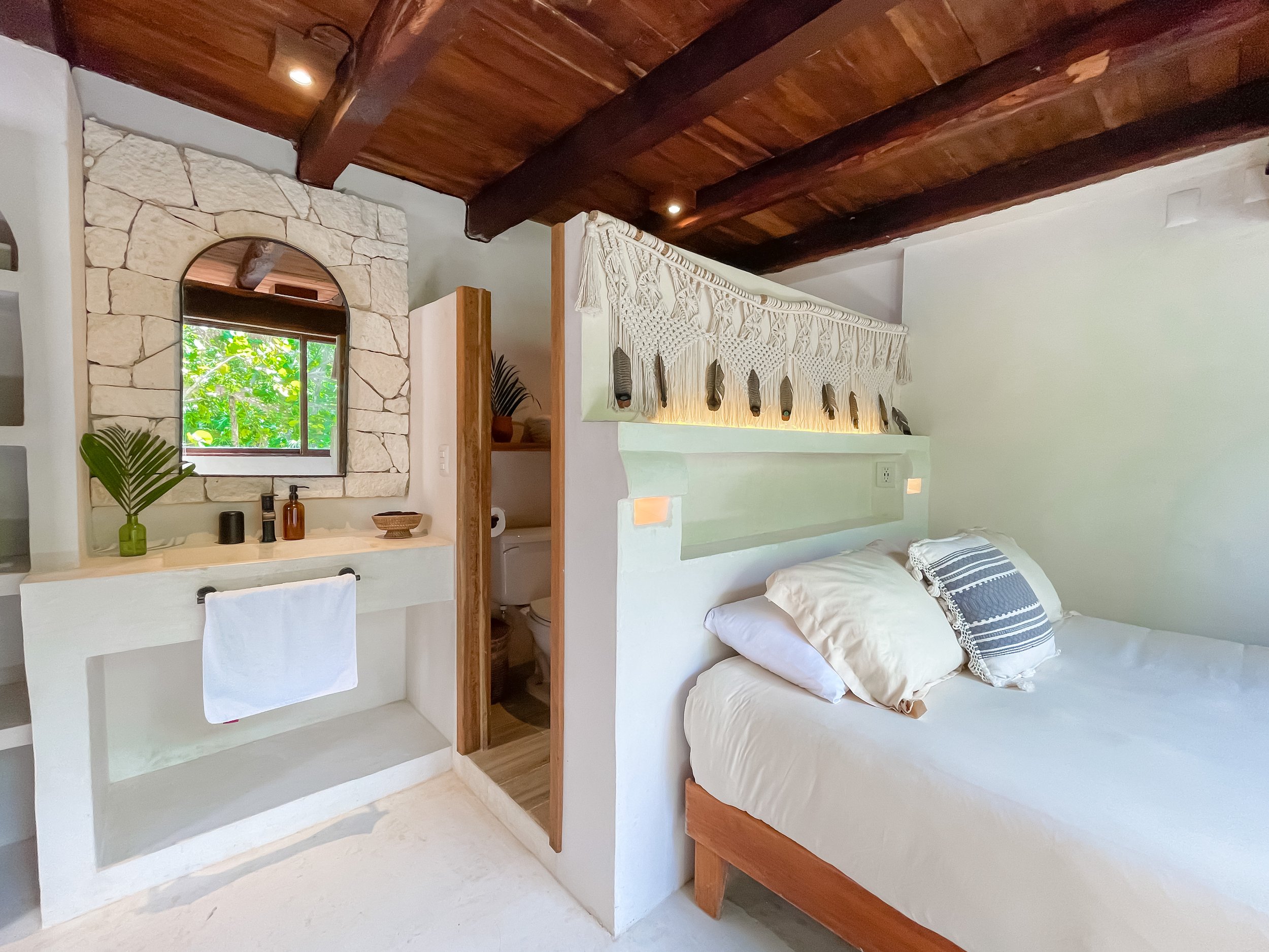 A cozy bedroom with a white bed and a patterned pillow. Behind the bed, there is a decorative wall hanging with tassels. To the left, a small vanity area with a mirror, candles, and a plant, and an open doorway leading to a bathroom with a toilet.