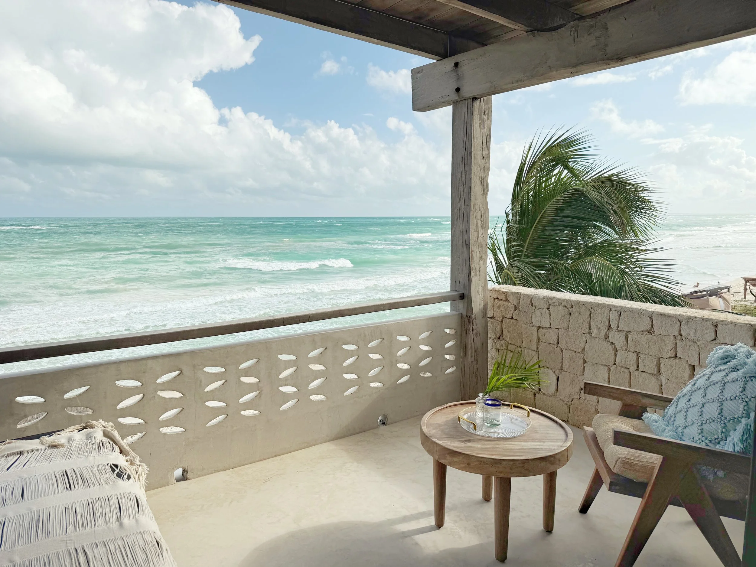 Beach view from a balcony with a small wooden table, a chair with a cushion, a blanket, a glass, a small plant, and a palm tree, overlooking the ocean with waves and a partly cloudy sky.