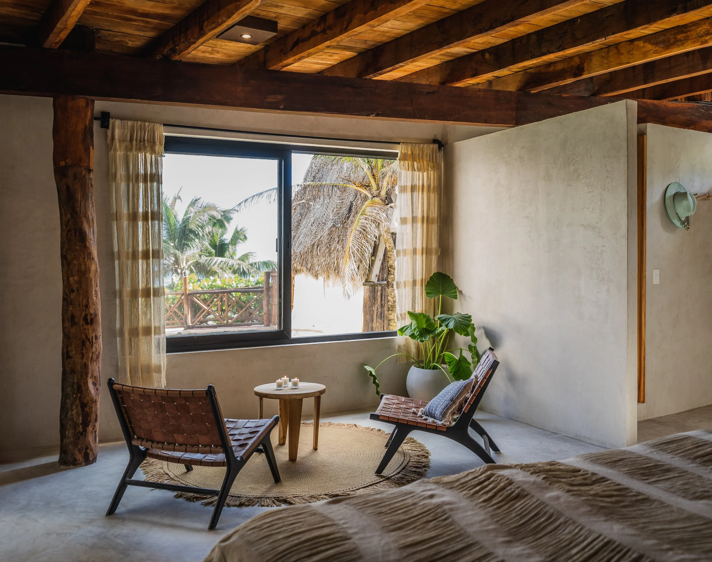 A cozy bedroom with a large window overlooking a tropical scene with palm trees and a thatched roof hut. The room features wooden beams, a small seating area with two chairs, a round table, a potted plant, and natural decor.