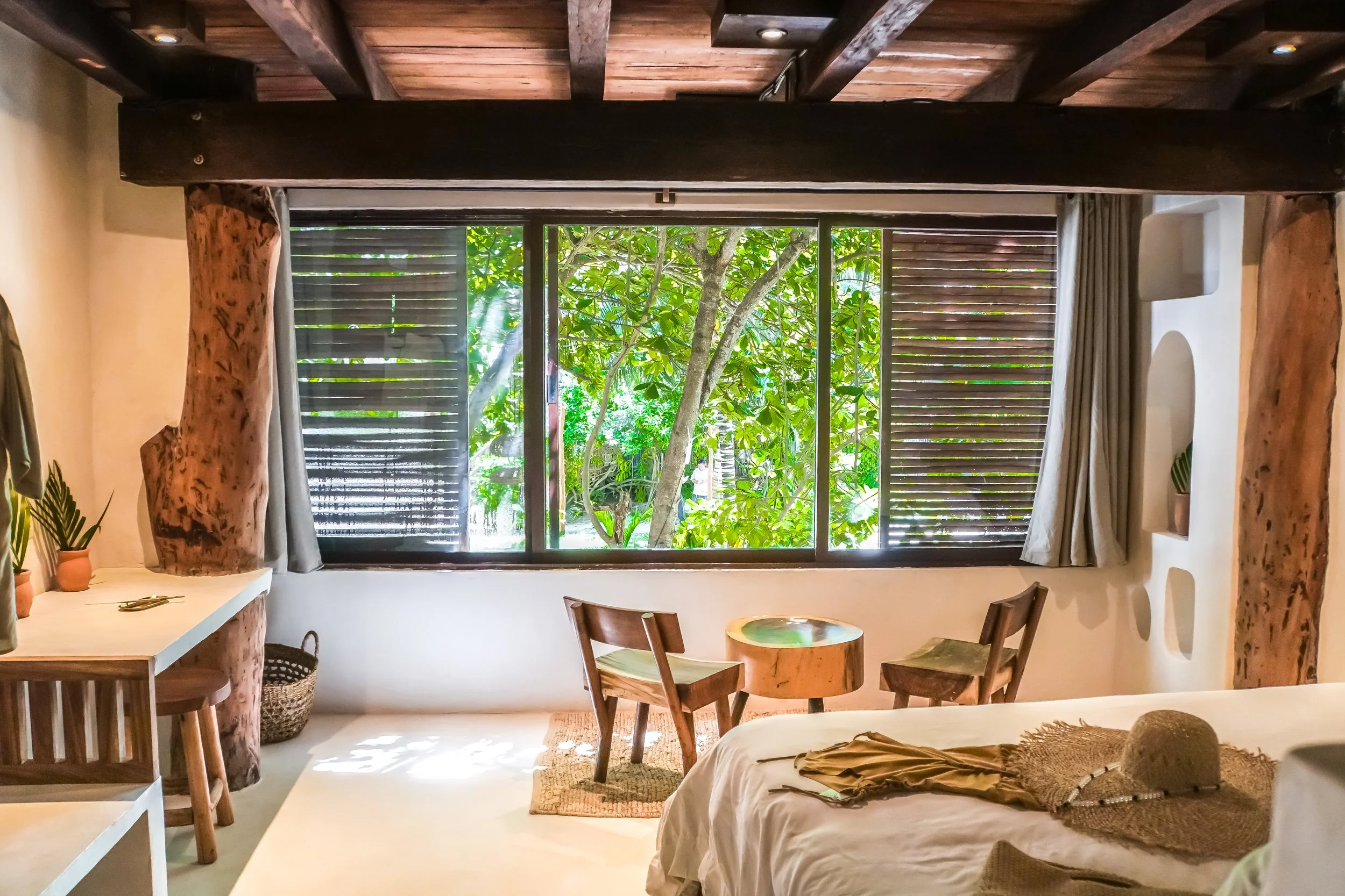 A cozy bedroom with a large window overlooking a lush green garden. The room has wooden beams on the ceiling, a white wall with built-in shelves, and a bed with a straw hat and dried plants on top. There are two wooden chairs, a small round table made from a tree trunk, and some plants in pots.