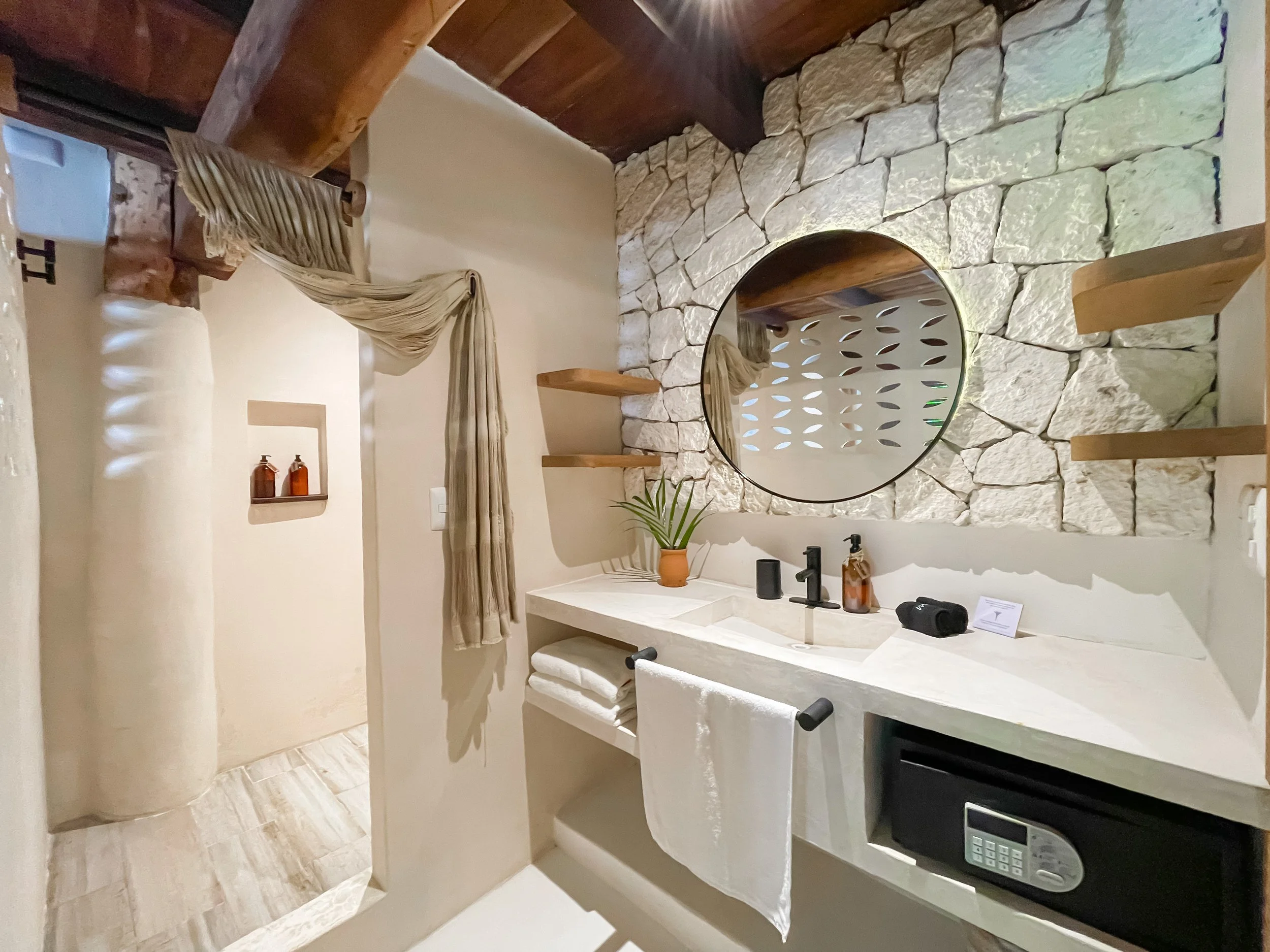 Modern bathroom with a stone accent wall, round mirror, potted plant, and a white vanity with towels, soap dispenser, and safe. Shower area with wooden beam and beige curtain.