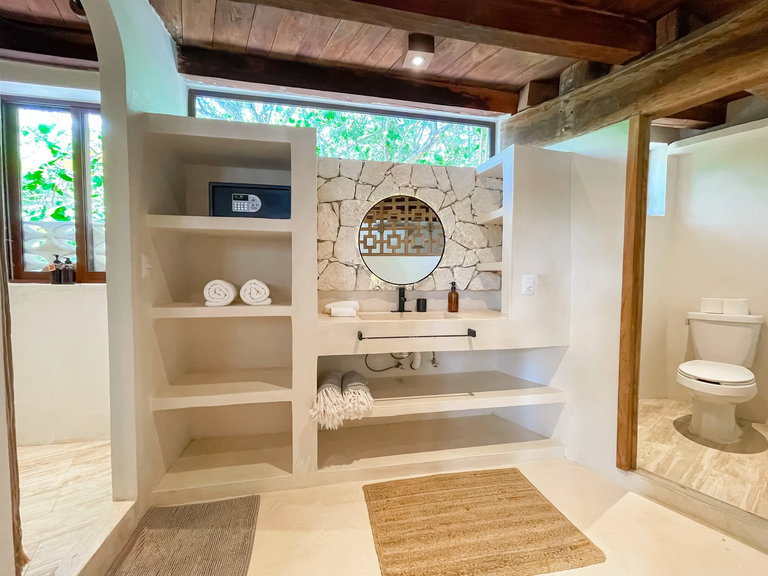 Bathroom with a stone accent wall behind the sink, a round mirror, a floating countertop with soap dispenser, and a window above showing greenery outside. To the right is a separate toilet area with wooden support beams across the ceiling.