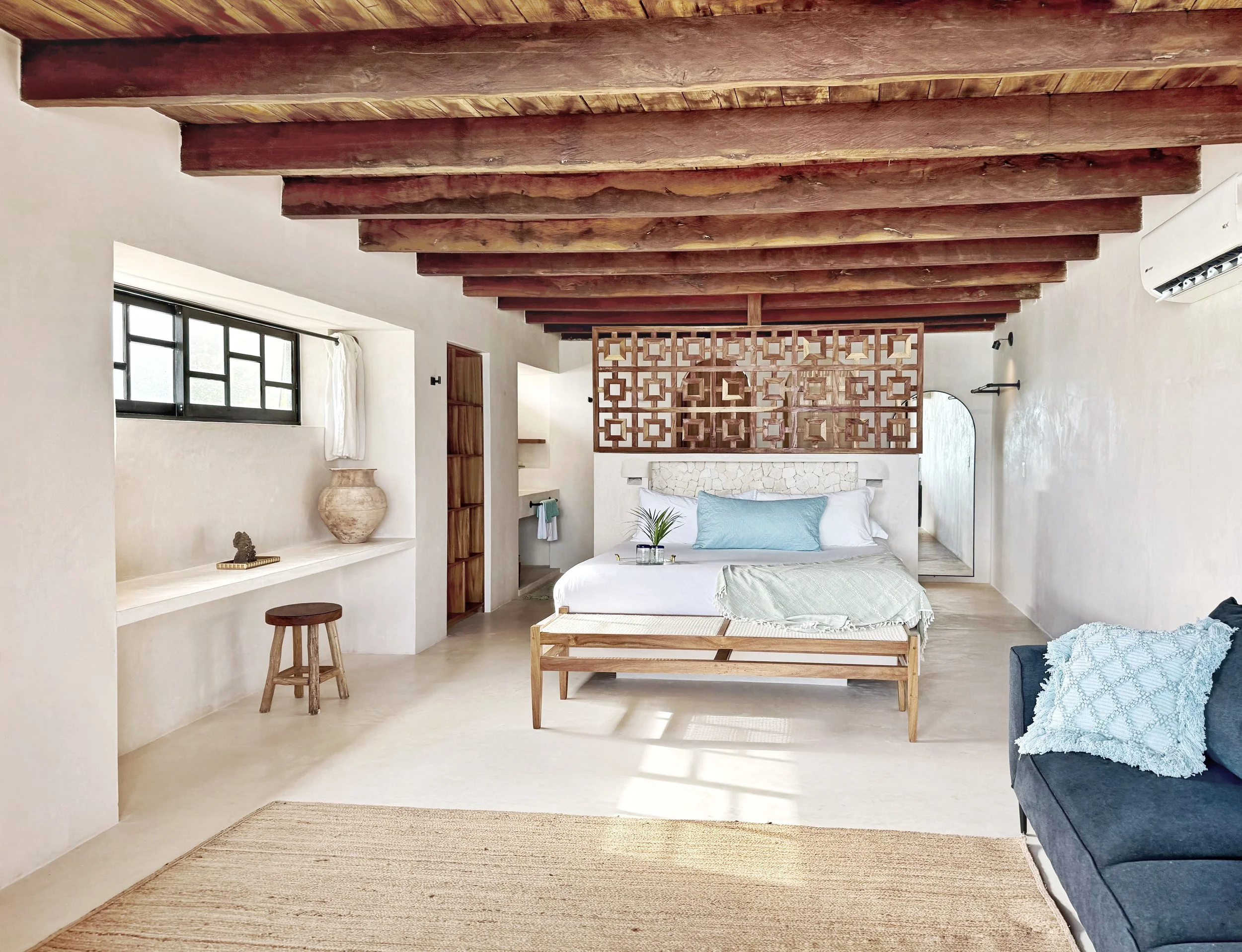 A bright bedroom with white walls, a wooden ceiling with exposed beams, a large bed with white and light blue pillows, a dark blue sofa with a textured white pillow, a built-in window ledge with a large ceramic vase, a small wooden stool, and a decorative wooden partition behind the bed.