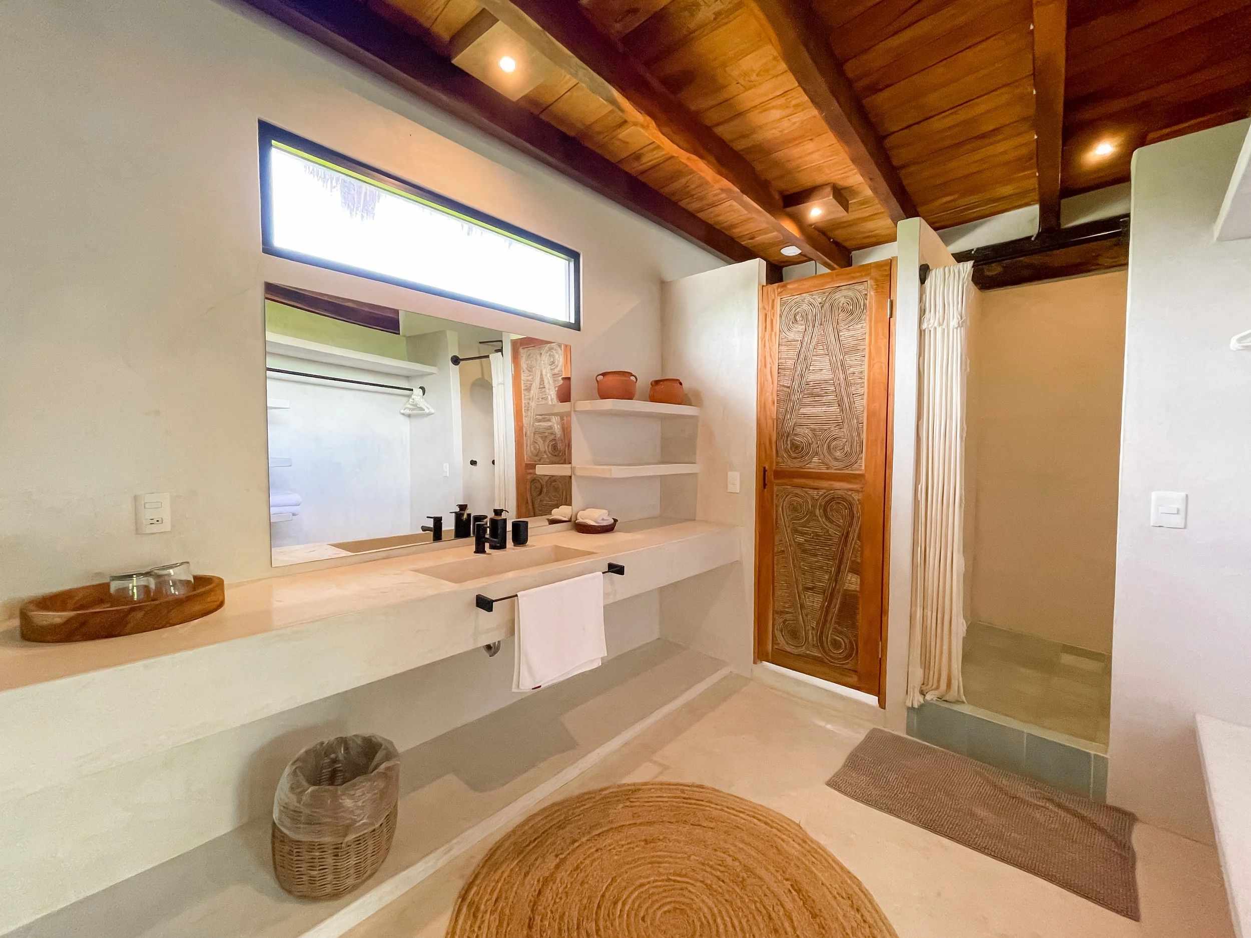 Bathroom with a large mirror, a cream-colored countertop with a black towel bar, black faucet fixtures, shelves with pottery, a wooden door with intricate carving, a shower area with curtain, and a woven circular rug on the floor.