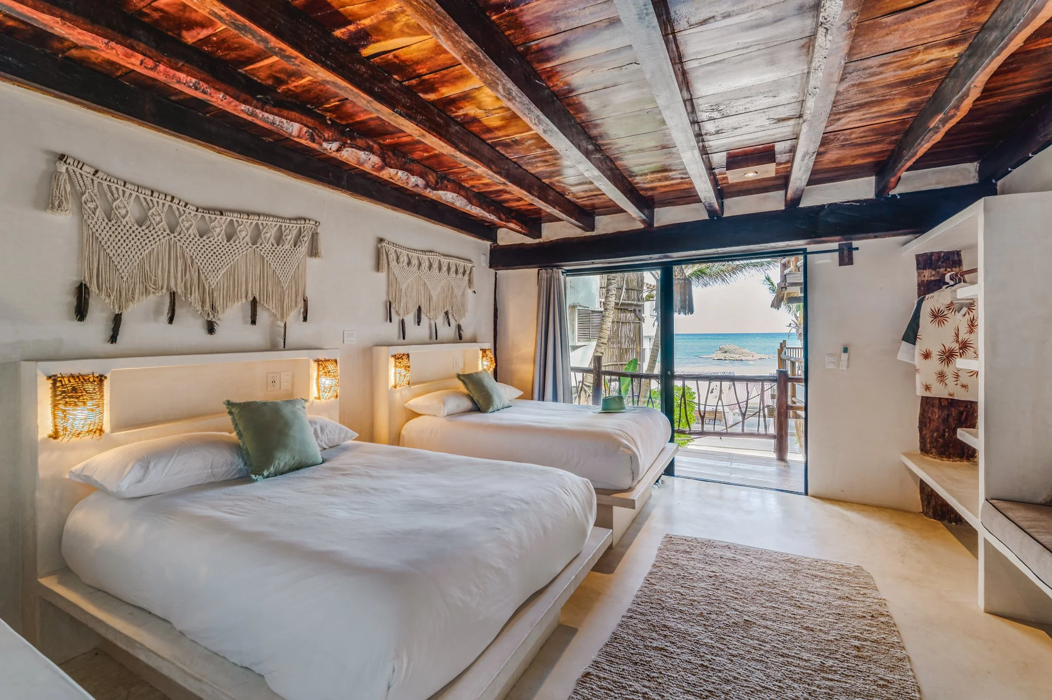 Beachside hotel room with two beds, decorative wall hangings, a sliding door opening to a balcony with ocean view, and tropical scenery outside.