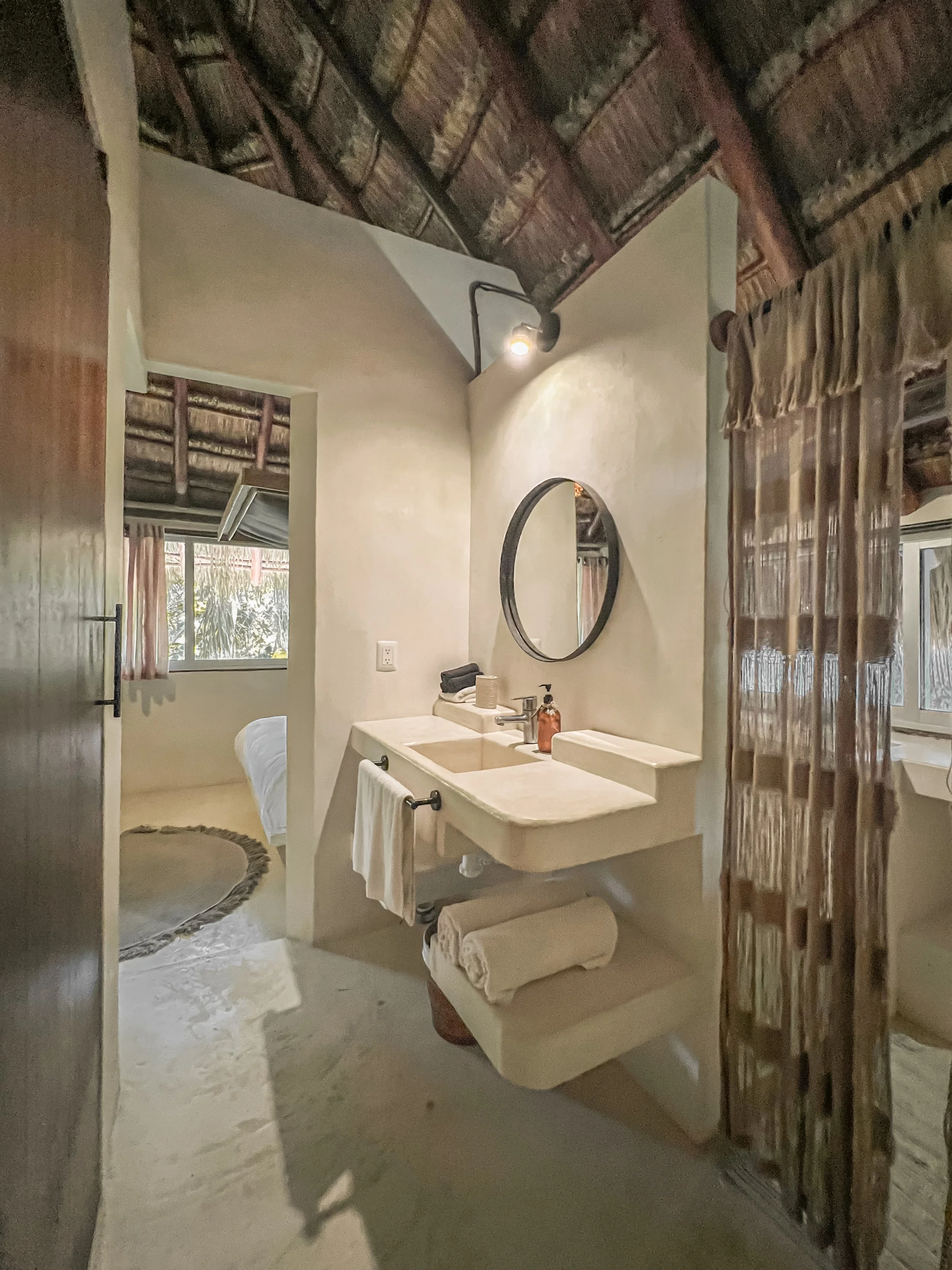 Bathroom with a stone sink, a round mirror, towels, soap dispenser, and a rustic curtain dividing the space, with a bed and window visible in the background.