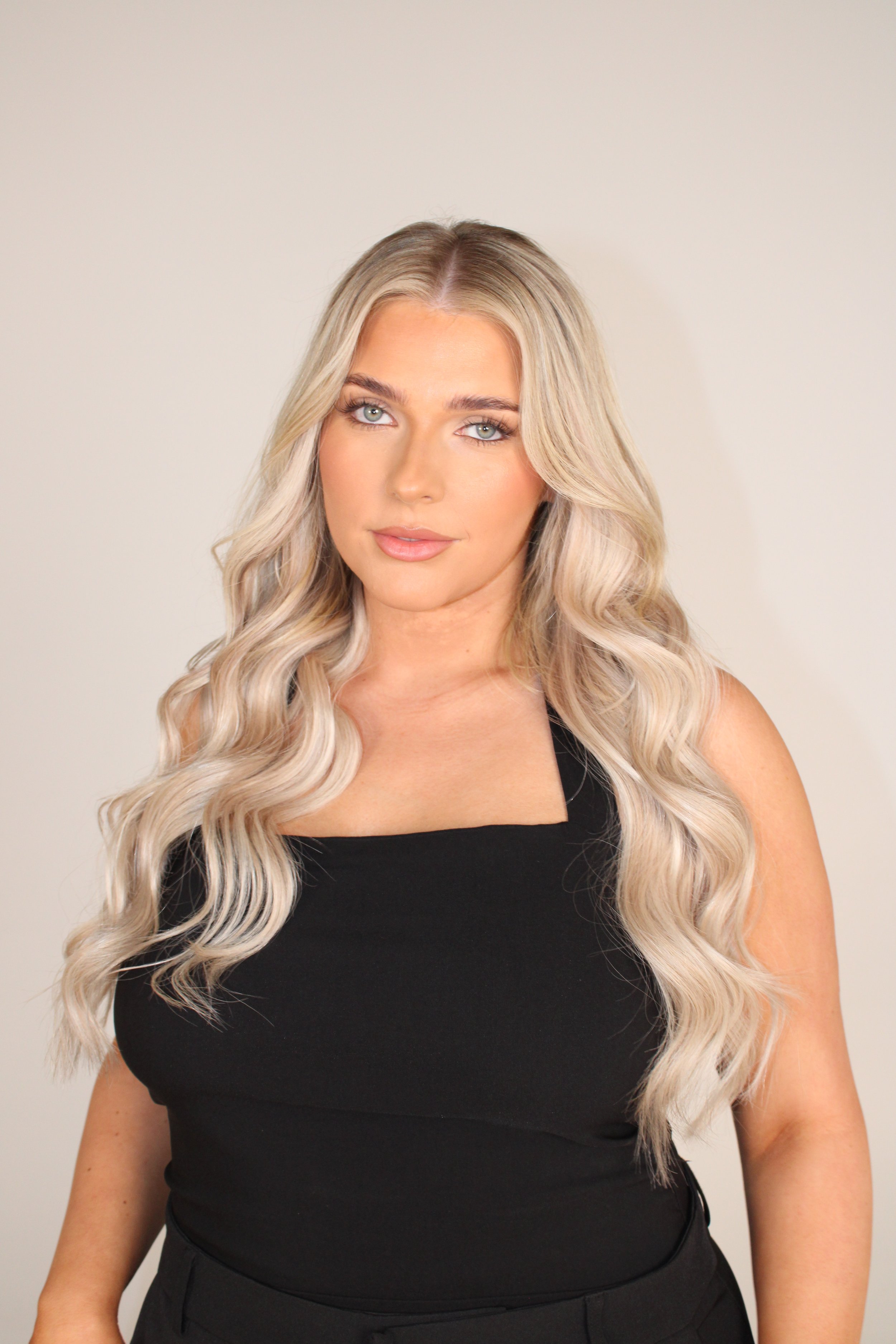 A woman with long, wavy blonde hair wearing a black top and black pants, standing against a plain light-colored background.