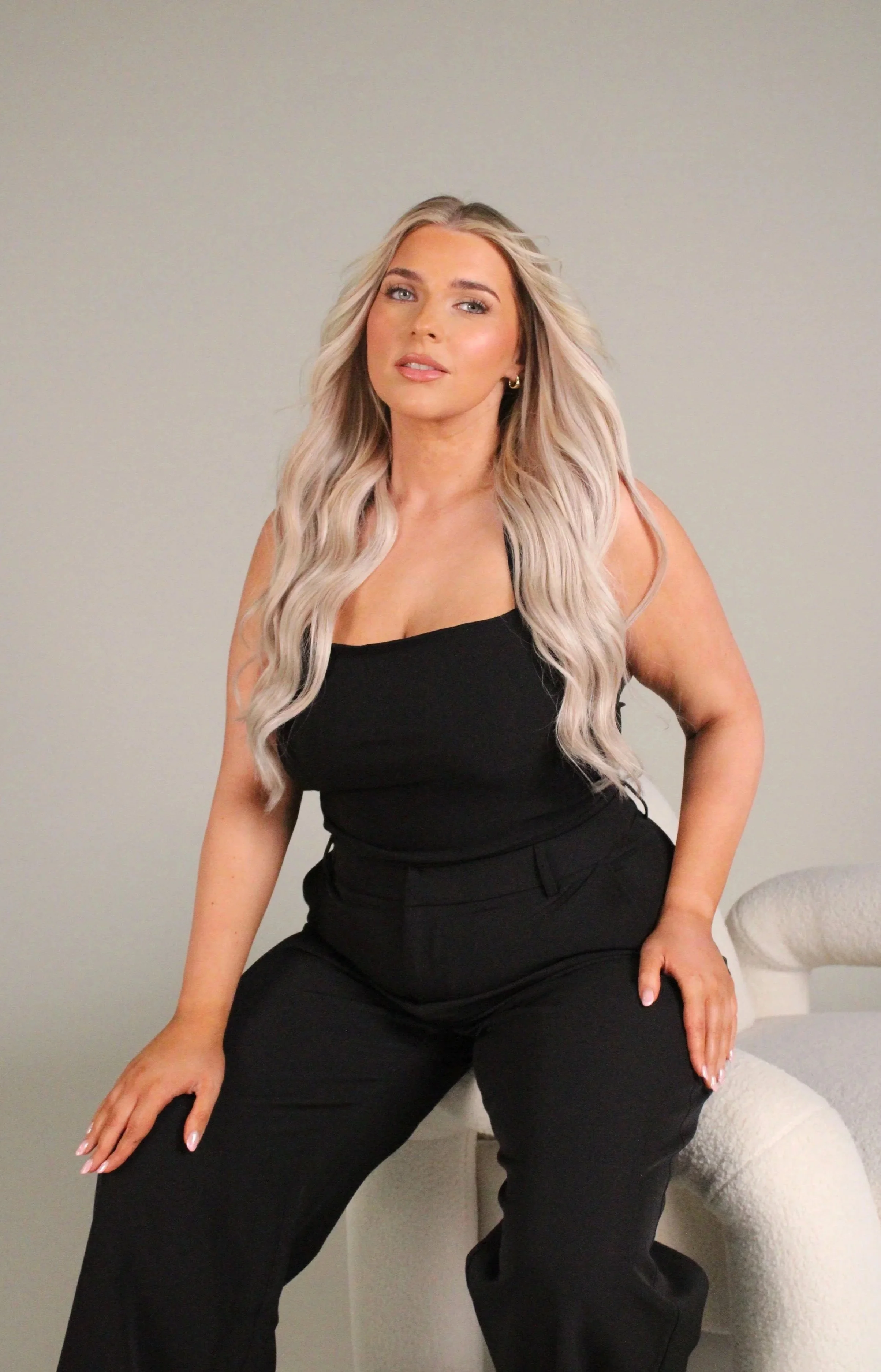 Woman with long wavy blonde hair wearing a black sleeveless top and black pants, sitting on a wicker chair in a room with light-colored walls.