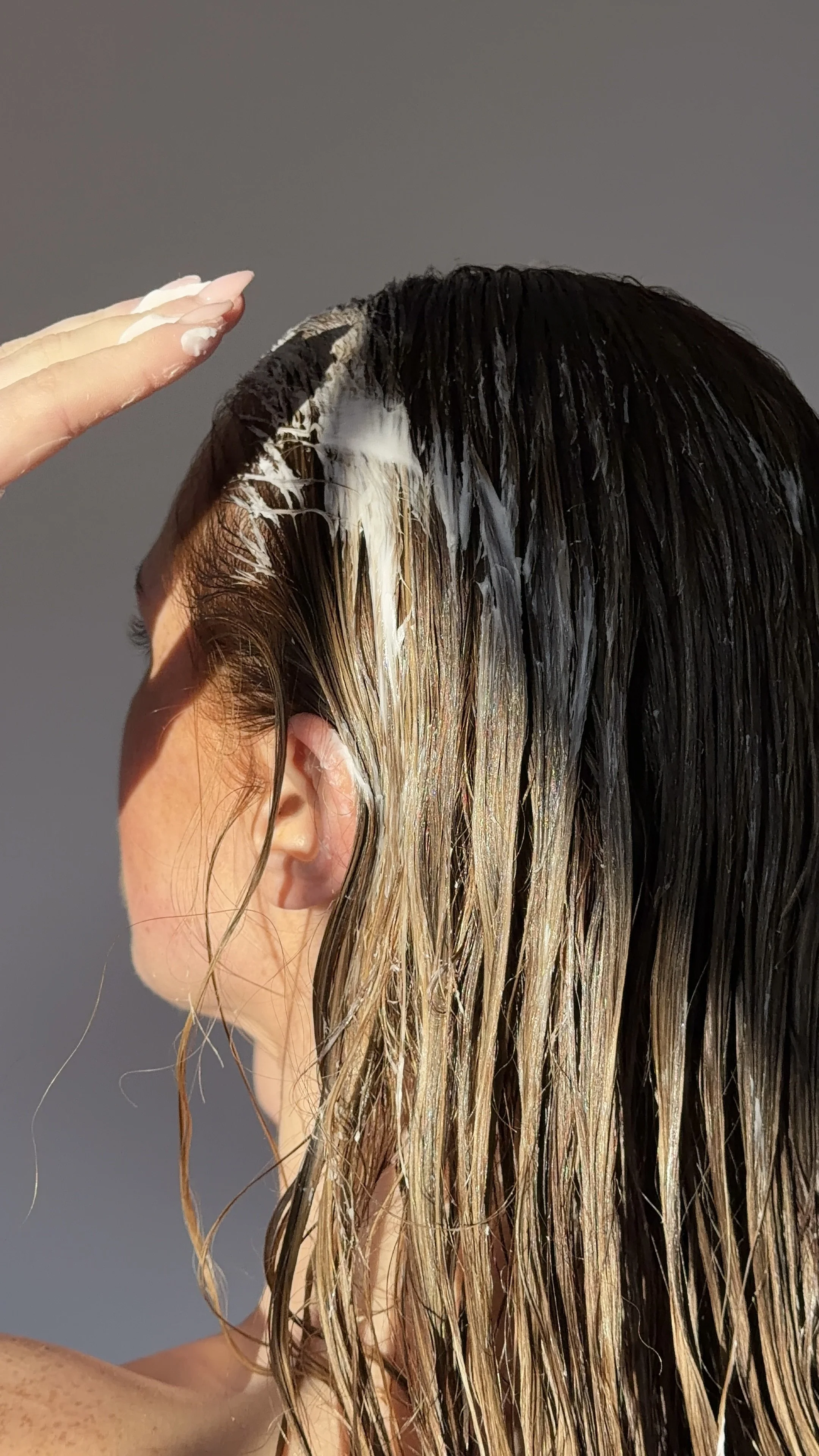 A person with blonde hair applying hair treatment or conditioner to their wet hair, which appears to be partially wet and styled in long strands, with a gray background.