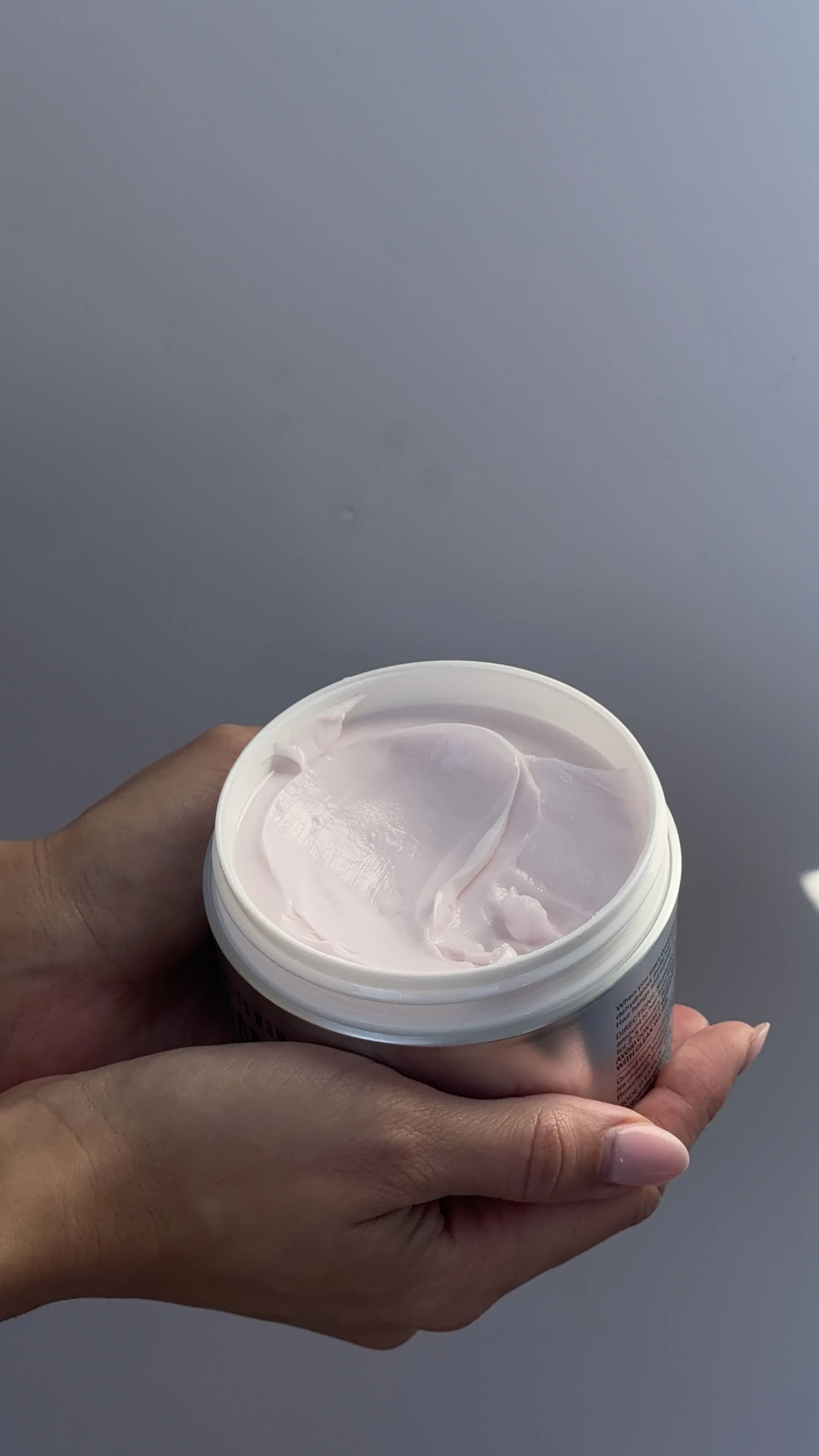 A person holding an open container of light pink cream or lotion against a gray background.