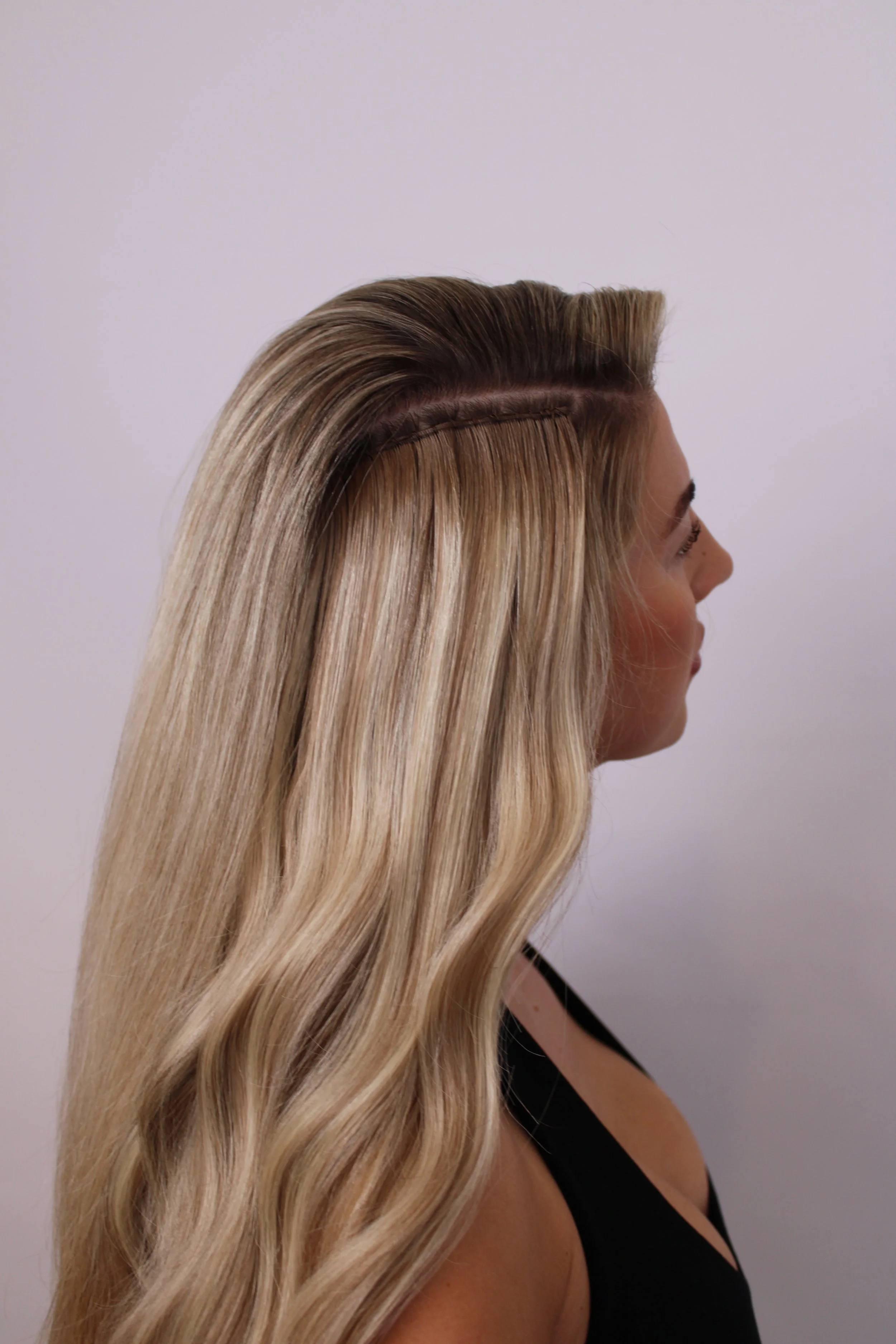 A woman with long, blonde hair styled in loose waves, wearing a black top, is seen in profile against a plain white background.