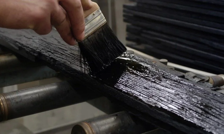 A person applying black paint or stain to a piece of wood using a brush.