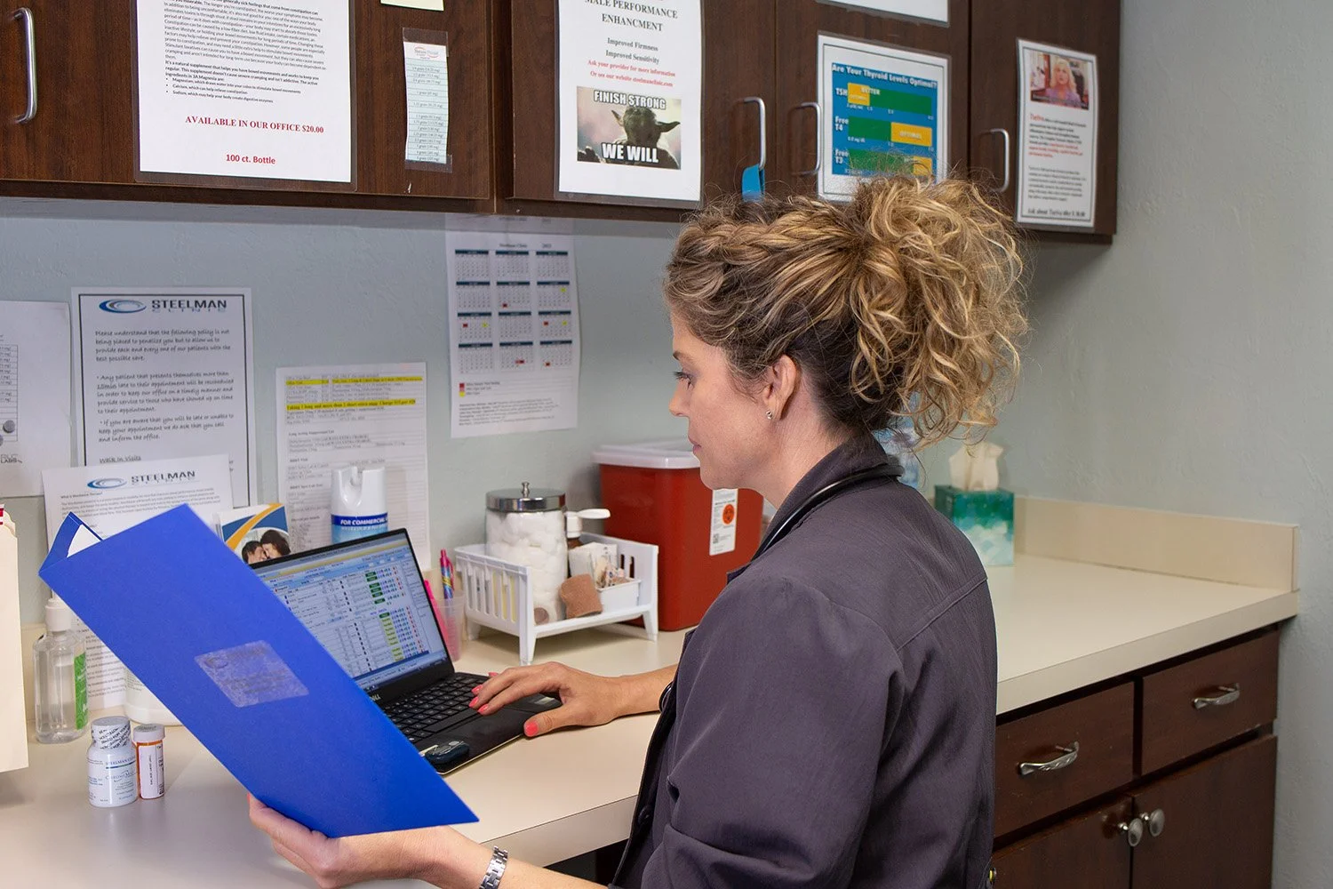 A woman with curly hair working on a laptop in a medical or office setting, holding a blue folder, with various documents, medical supplies, and informational posters on the wall behind her.