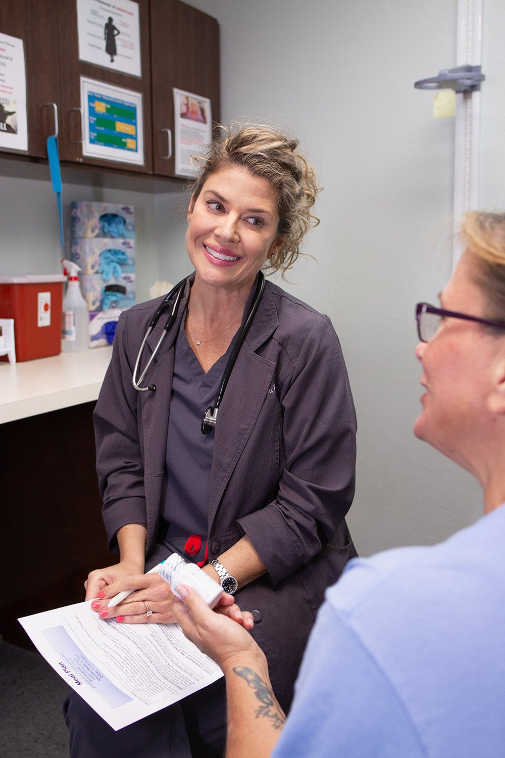 A female healthcare professional with a stethoscope around her neck and a patient sitting across from her, holding a medication bottle and a script in a medical office.