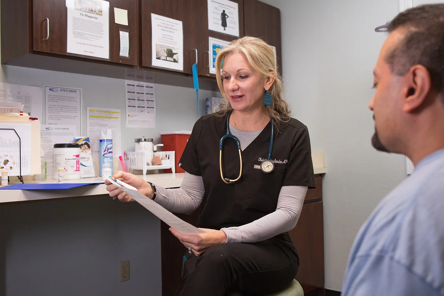 A female healthcare professional with a stethoscope around her neck is sitting and reading a document to a male patient in a medical office or clinic.