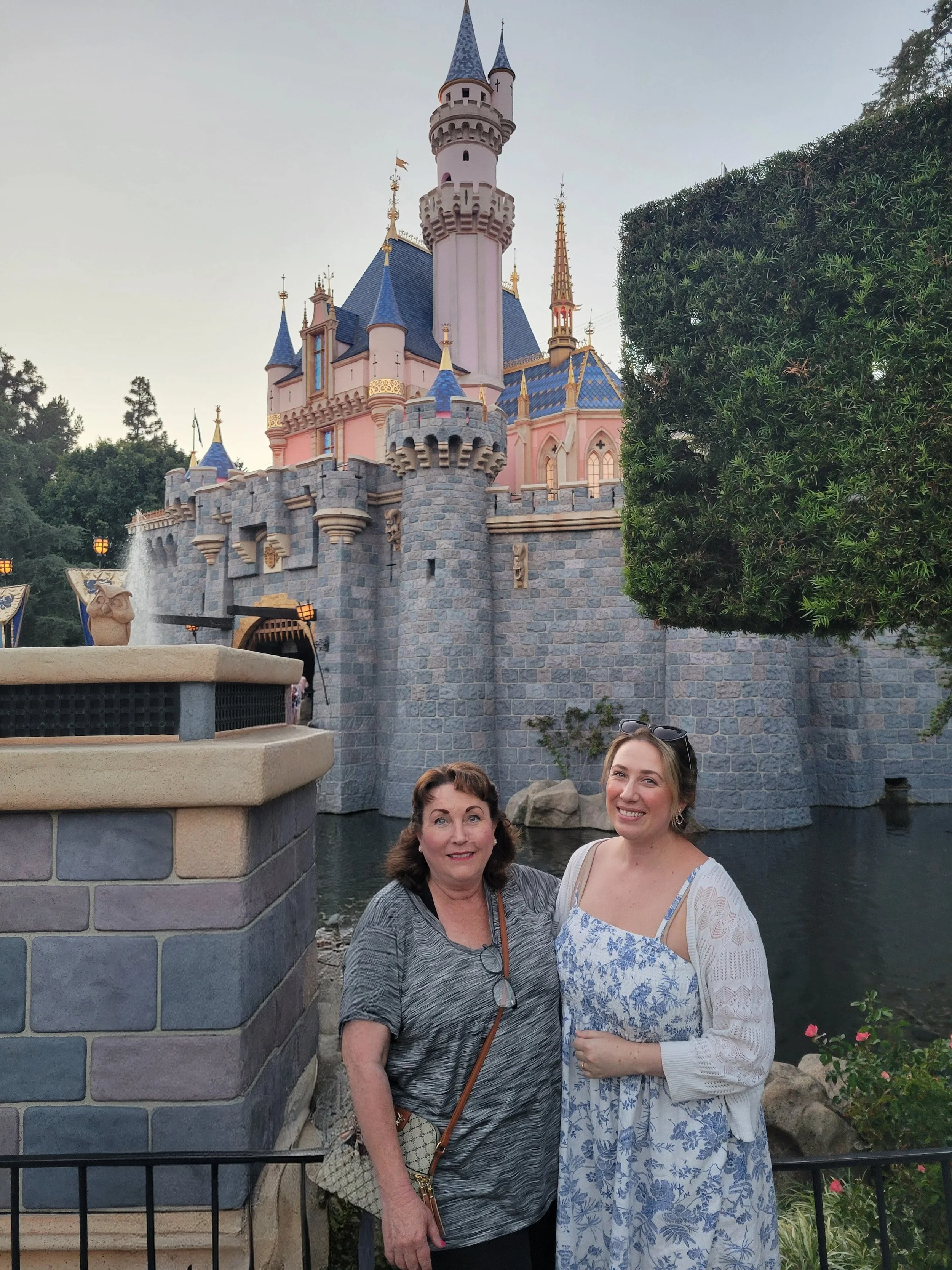 Two women standing in front of Sleeping Beauty Castle at Disneyland, with the castle in the background and a large shrub on the right side of the image.