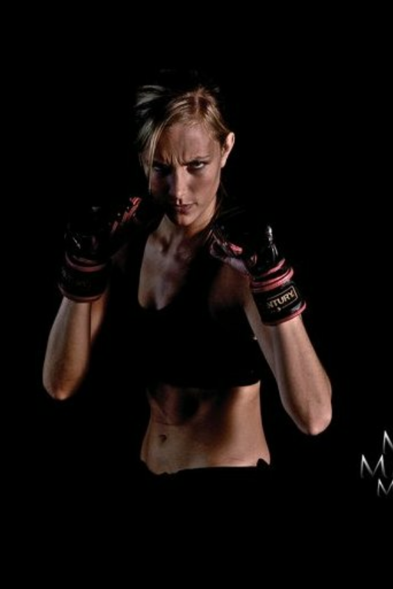 A female boxer in a fighting stance with gloves, wearing a black sports bra, and looking intensely at the camera against a dark background.