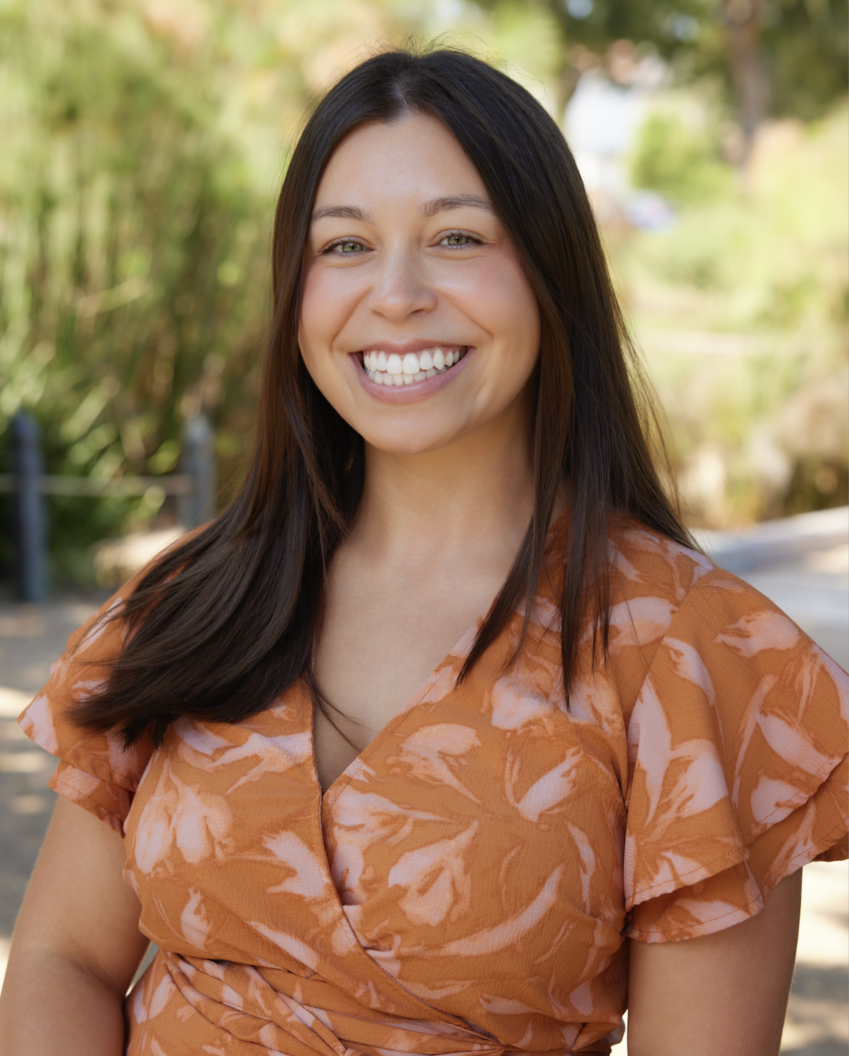 A smiling woman with long dark hair, wearing an orange patterned top, outdoors with trees and sunlight in the background.
