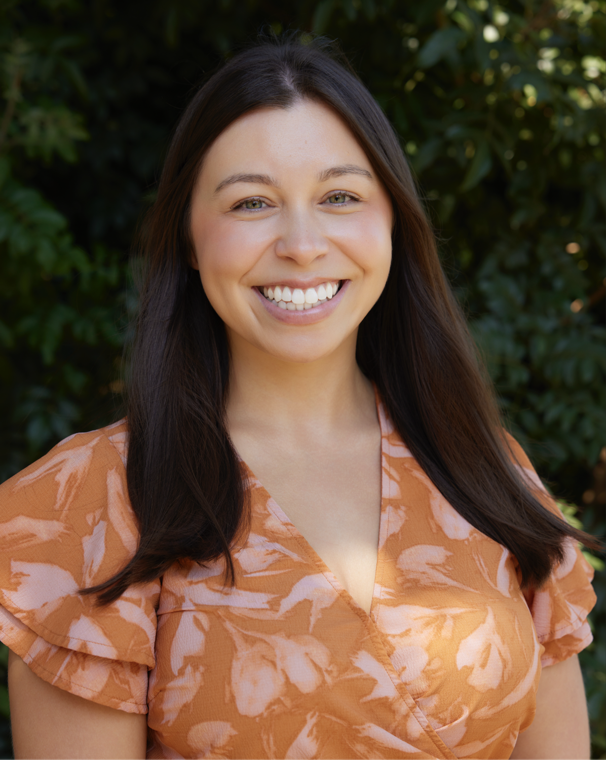 A smiling woman with long dark hair standing outdoors with green foliage in the background, wearing an orange patterned blouse.