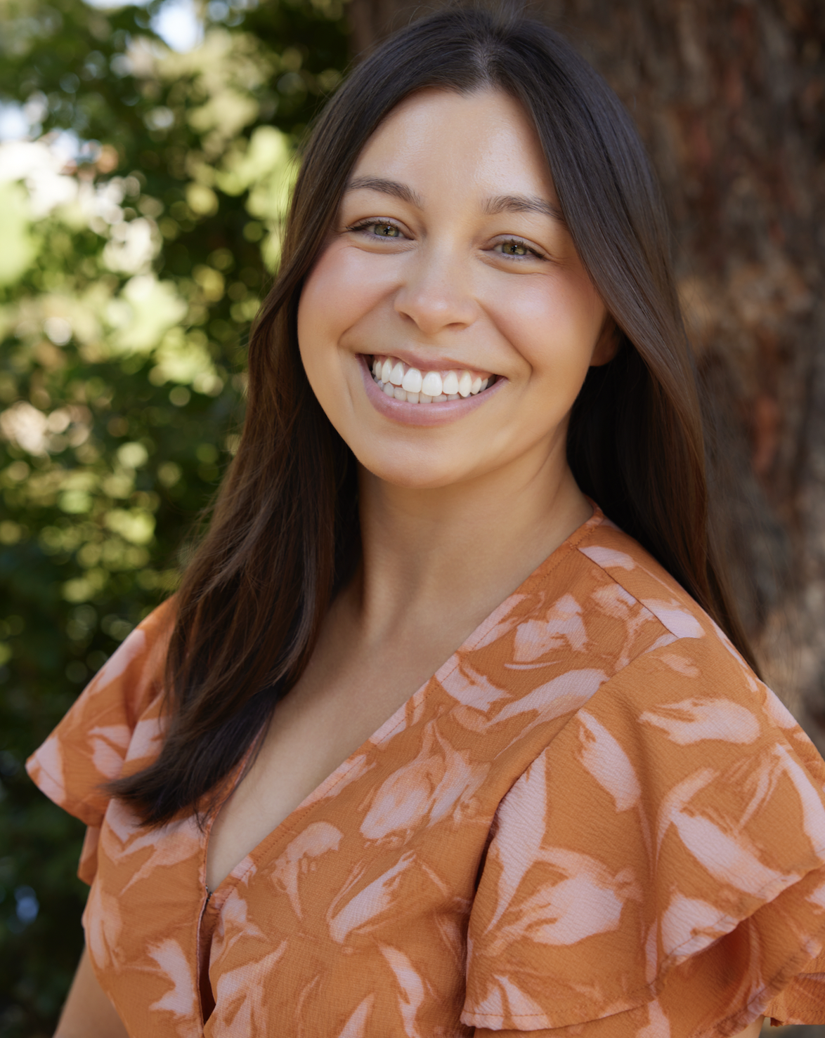 A young woman with long brown hair smiling outdoors with a tree and greenery in the background, wearing an orange patterned blouse.