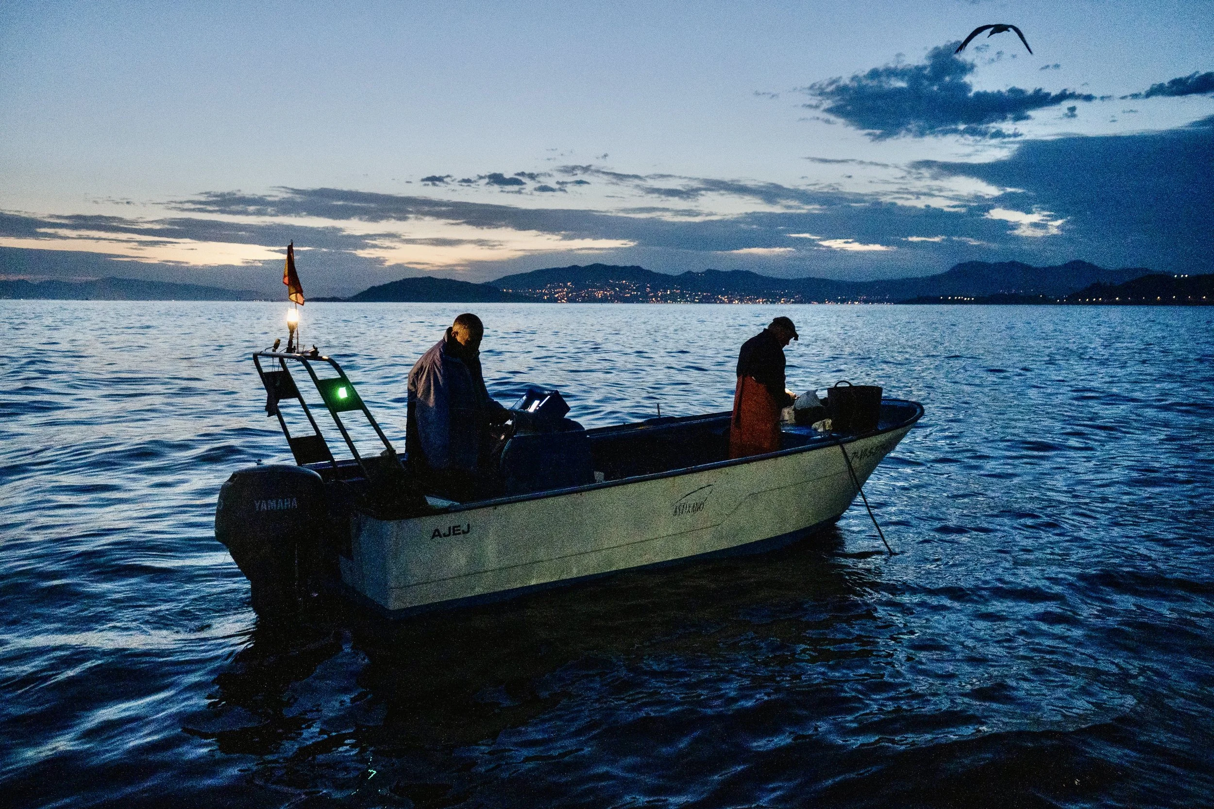 Dos personas en un bote en un lago al atardecer, con montañas y ciudad al fondo.