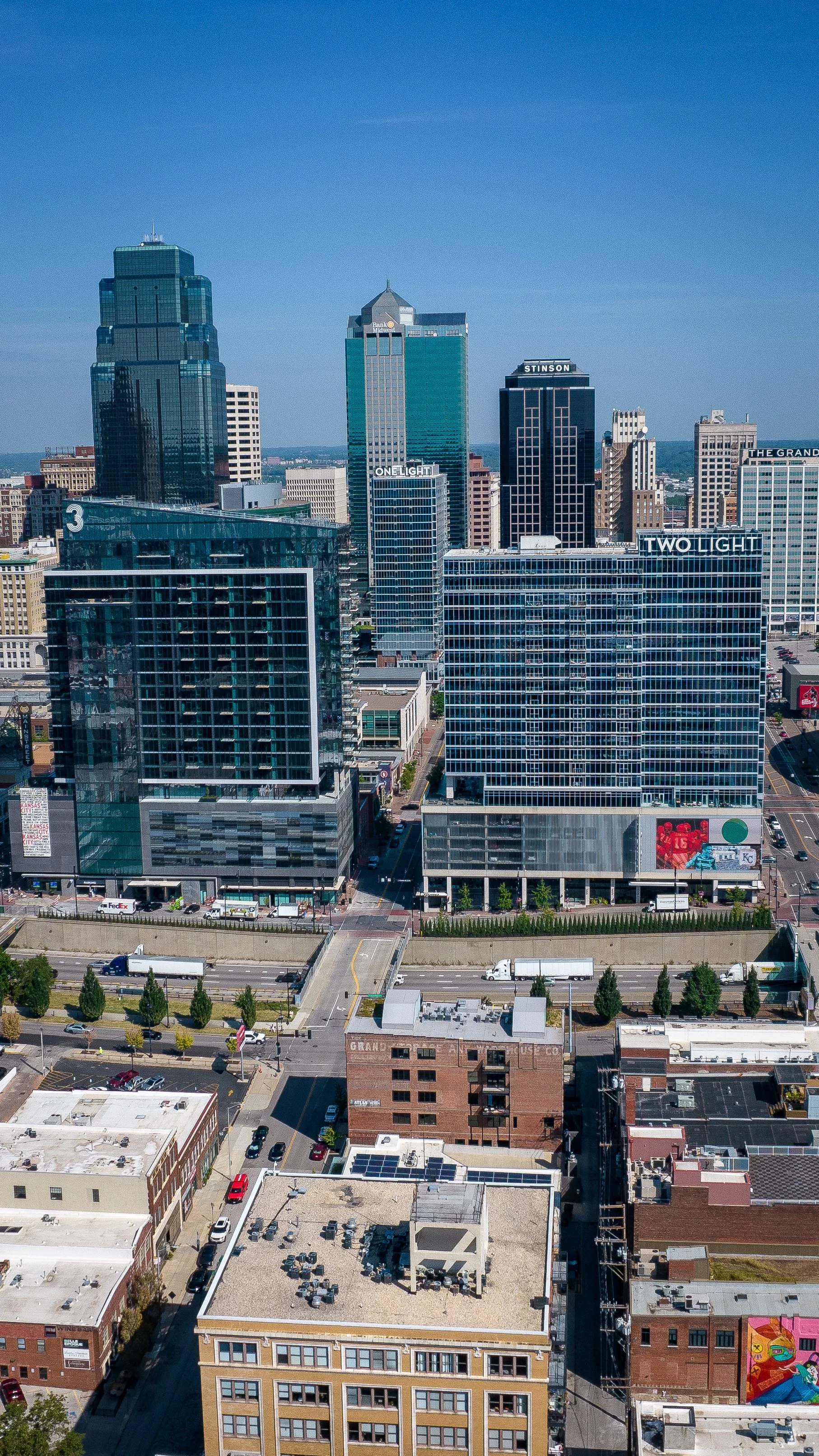 A city skyline with tall modern skyscrapers and office buildings, including signs for 'TWO LIGHT', 'ONE LIGHT', and 'STINSON', under a clear blue sky, with streets, trees, and smaller buildings in the foreground.