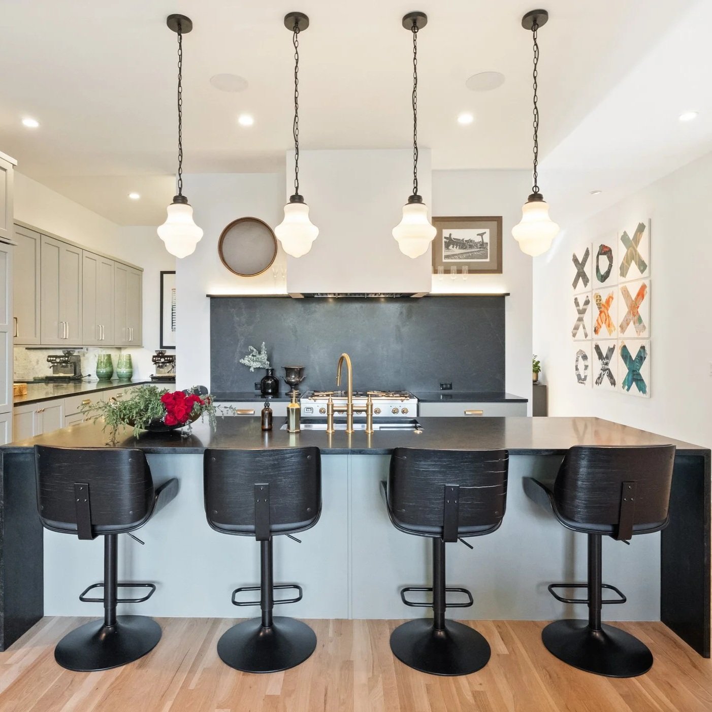 Modern kitchen with central island, black countertop, four black bar stools, white cabinetry, black backsplash, gold fixtures, pendant lights, wall art, and decorative items.