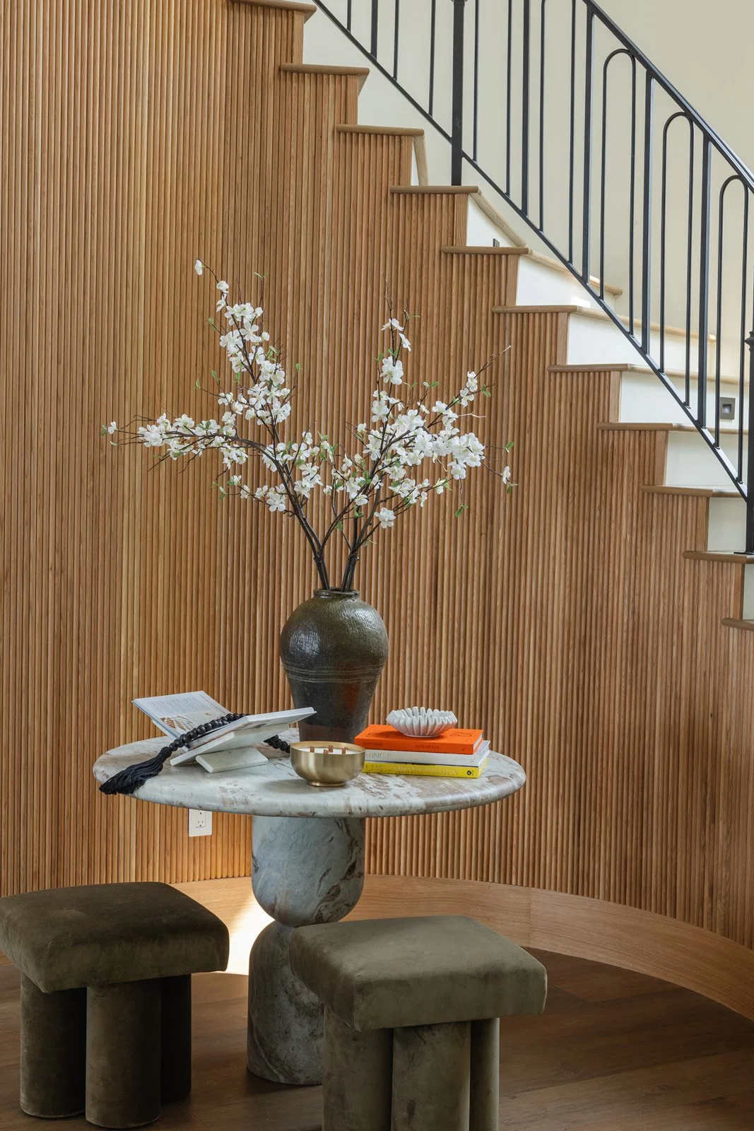 A marble table with a large vase of white flowers, books, and decorative items, with wooden wall paneling and a staircase in the background.