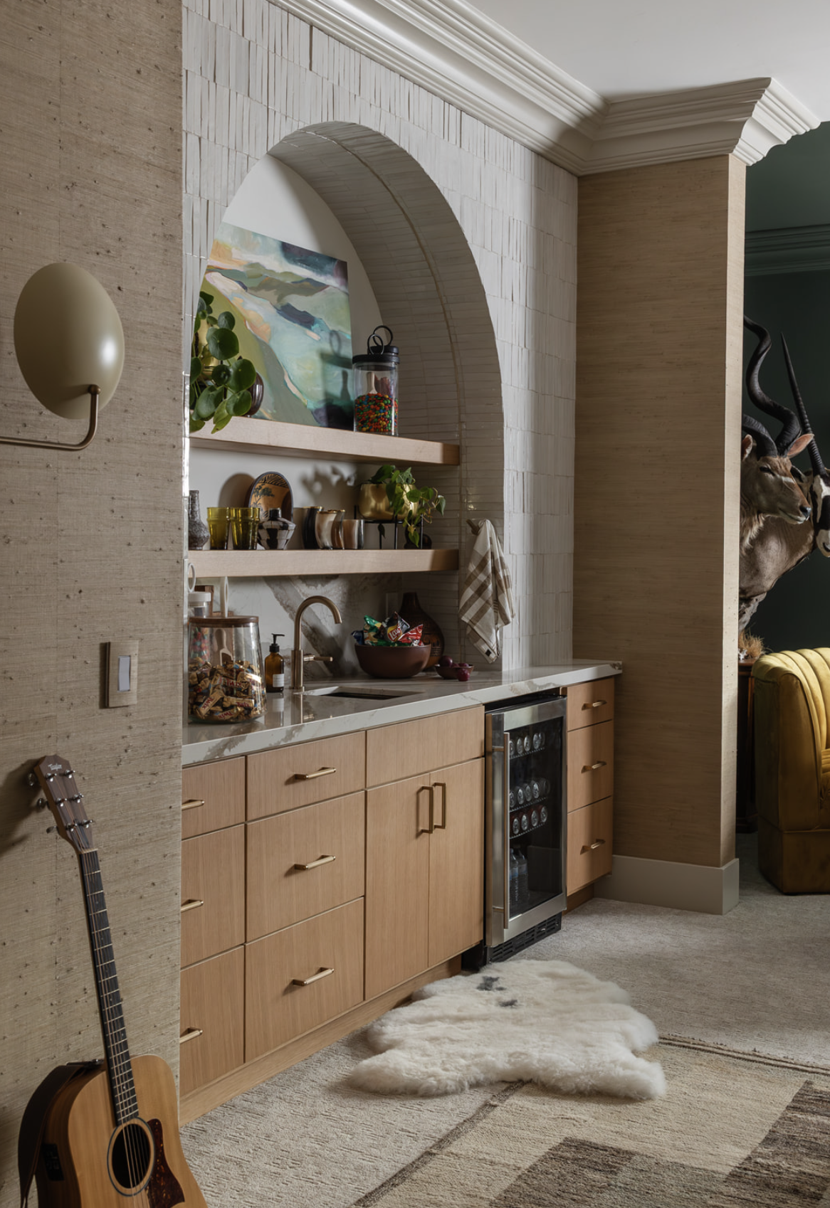 Interior view of a modern kitchen with beige cabinets, a white countertop, and open shelves decorated with plants, books, and bowls. An arched wall niche with art and jars. A small wine fridge is built into the cabinetry, and a white sheepskin rug li
