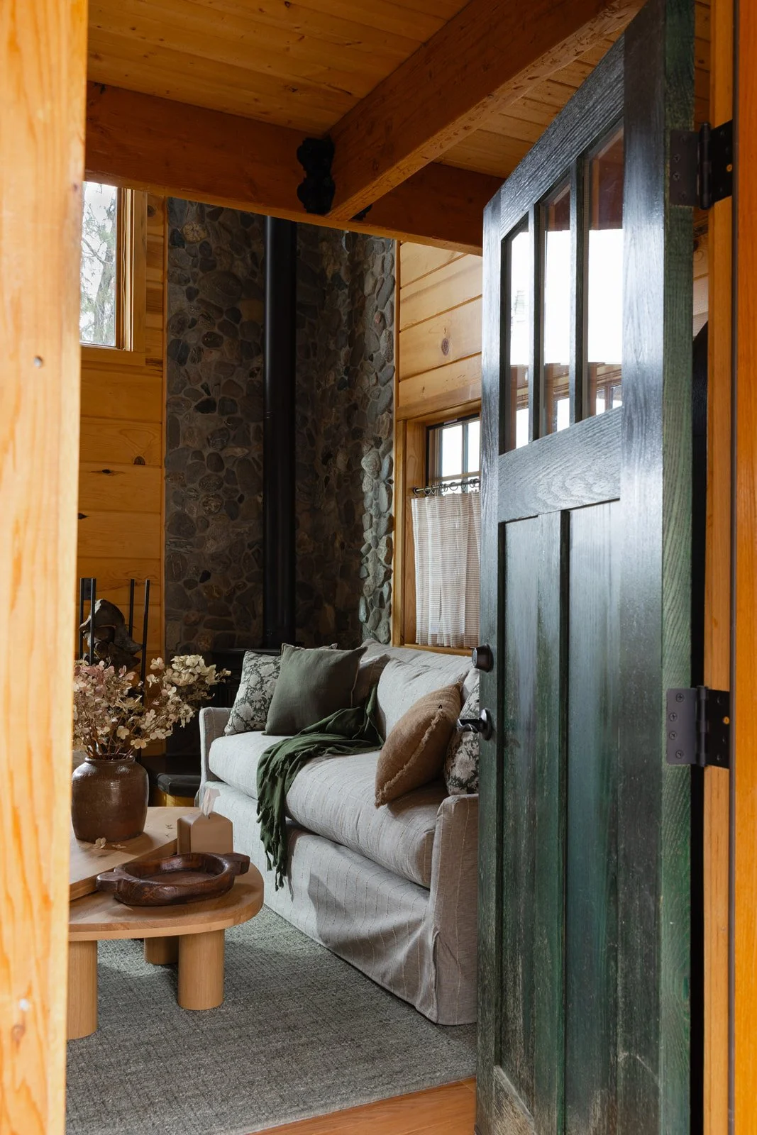 A cozy living room with a beige sofa, green and beige pillows, and a green throw blanket, viewed through a partly open dark green wooden door. There are wedged windows with curtains, a stone wall around a black stove pipe, and a wooden ceiling with b